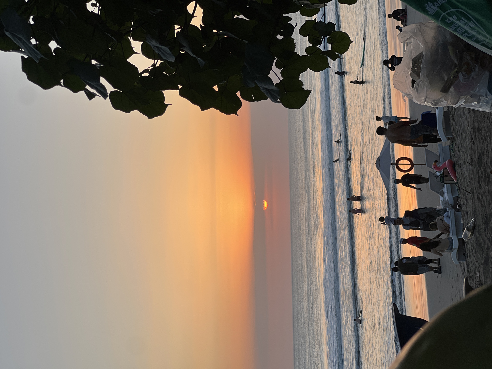 This is a photograph taken at sunset on the beach in Kuta, Indonesia. The sun is low on the horizon, casting a warm orange glow across the sky and the ocean. Gentle waves roll onto the sandy shore. Several people are visible on the beach and in the water. Some are wading, others are standing on the shore, and a few are in the water with surfboards. In the foreground, silhouetted against the sunset, are several lounge chairs, an umbrella, and a trash bag filled with items. To the right, a leafy tree branch hangs over the scene, also silhouetted. The overall impression is one of a tranquil beach scene at dusk.