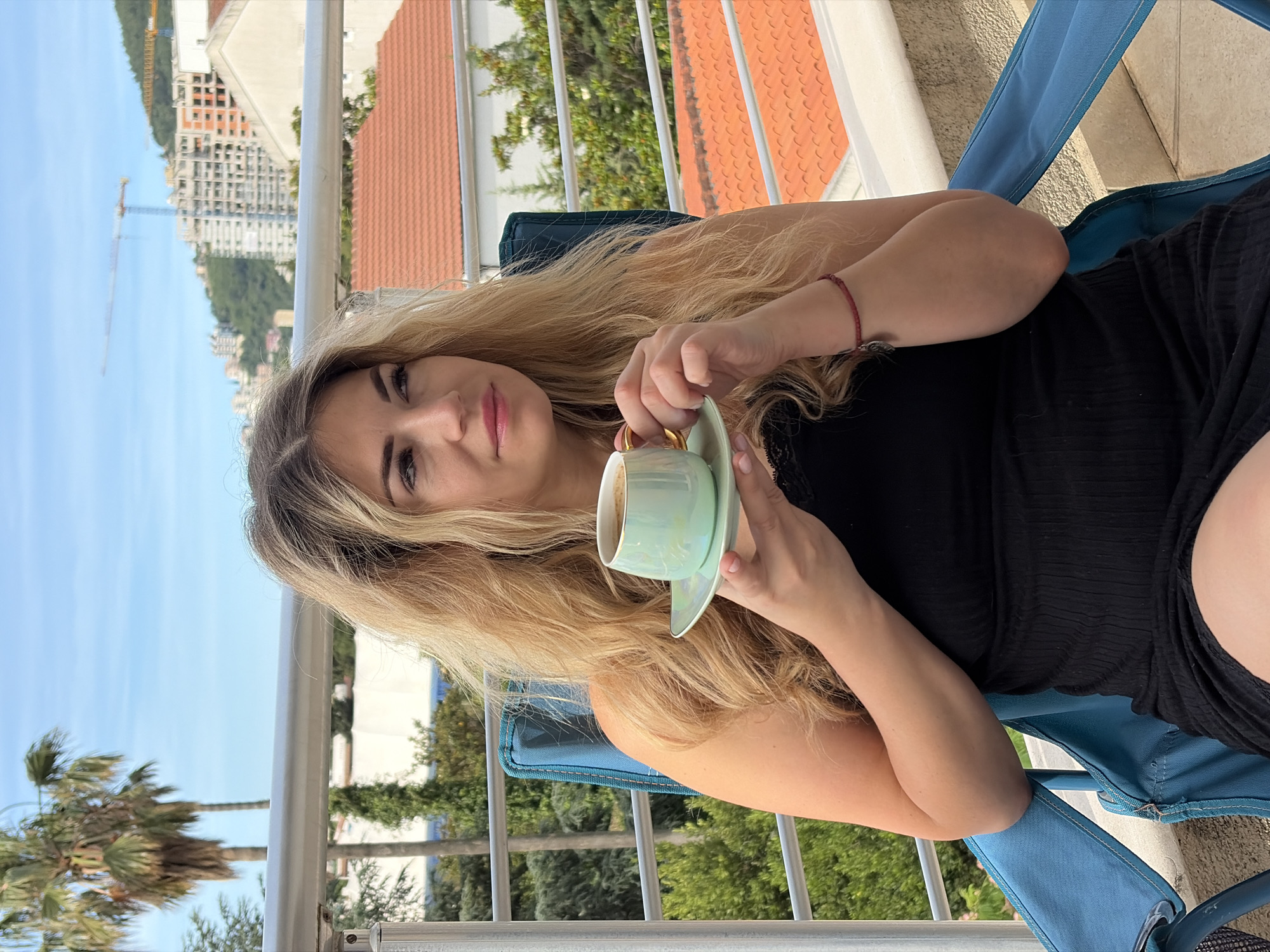 A young woman with long, light blonde hair is seated in a blue outdoor chair on a balcony, looking directly towards the viewer with a relaxed expression. She is wearing a black top or dress and has a red bracelet on her left wrist. In her hands, she delicately holds a light blue or mint green teacup with a matching saucer, both featuring gold trim. The cup appears to contain a beverage like coffee.
The setting is a sunny outdoor environment, likely a balcony, indicated by a metal railing that spans the foreground. The background reveals a panoramic view of a city, consistent with Budva, Montenegro, under a clear blue sky. On the left side of the frame, distant mountains are visible, along with several large buildings under construction, characterized by their concrete frames and a prominent construction crane. A palm tree and other greenery are also visible. To the right, the cityscape includes buildings with reddish-orange tiled roofs and dense green foliage, suggesting a mix of developed and natural landscapes. The overall impression is one of a calm, leisurely moment on a bright day. No visible text is present in the image.