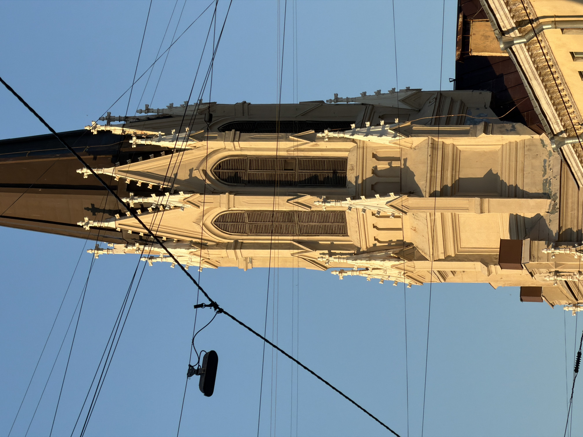 The image shows the upper section of a Gothic Revival style church tower in St. Petersburg, Russia. The tower is cream-colored with ornate white decorative elements, including pinnacles and arched windows with dark shutters. Several power lines and a street lamp are visible in the foreground, crisscrossing the sky and partially obscuring the tower. The sky is a clear, bright blue, indicating a sunny day. The lighting suggests the photograph was taken during the late afternoon or early morning, as indicated by the long shadows cast on the tower. The lower part of the image shows the tiled roof and upper floors of an adjacent building, also in a warm yellow hue.