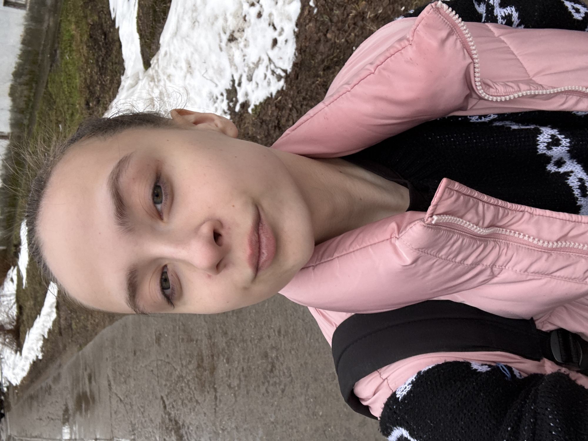 A young woman is outdoors on what appears to be a sidewalk or paved area, in the city of Chaikovsky, Russia. Patches of snow and bare ground are visible in the background. She is wearing a pink padded jacket over a black sweater with a white snowflake pattern. The camera's angle suggests the photo was taken at a slightly downward perspective.