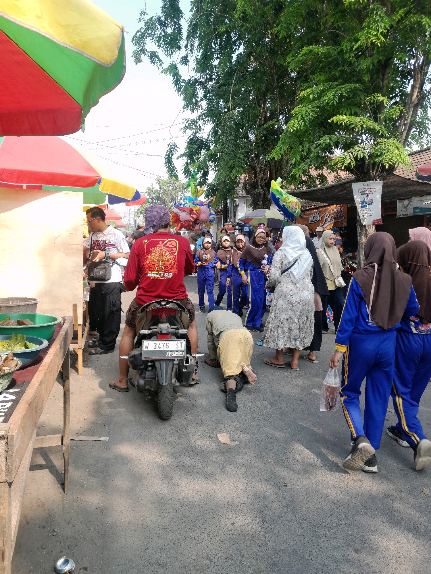 In the city of Jogosatru, Indonesia, on a sunlit day, a street scene unfolds. A person wearing a red long-sleeved shirt with elaborate yellow and red graphics on the back is seated on a motorcycle. The motorcycle's license plate displays "W 3476 ST". Beside the motorcycle, a person in a yellow garment is kneeling on the street, facing away from the camera. Several individuals in blue uniforms with yellow stripes are walking on the right side of the frame, some carrying plastic bags. Another person in a patterned dress and a white headscarf is also walking. In the background, colorful umbrellas are set up, possibly for vendors. Trees with lush green foliage line the street, and buildings with tiled roofs are visible. A table with green bowls containing food items is positioned on the left side of the image. The street is paved with asphalt, and shadows are cast across it by the umbrellas and trees. A balloon seller appears to be present further down the street, with a cluster of colorful balloons visible.