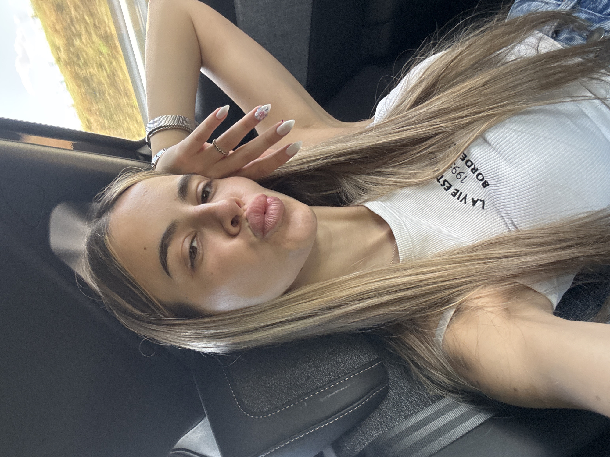 A young woman with long, blonde hair is taking a selfie inside a vehicle. She is wearing a white ribbed tank top with black text that reads "LA VIE EST 1998 BORDE". Her right arm is bent, resting on her head, and she is wearing a silver bracelet and a ring on her left hand. Her lips are pursed, as if she is blowing a kiss. The background shows the interior of a car, with a dark seat and a window showing a blurred landscape of a highway and a grassy hill. The lighting suggests it is daytime. The scene does not appear to be in Zheleznik, Bulgaria.