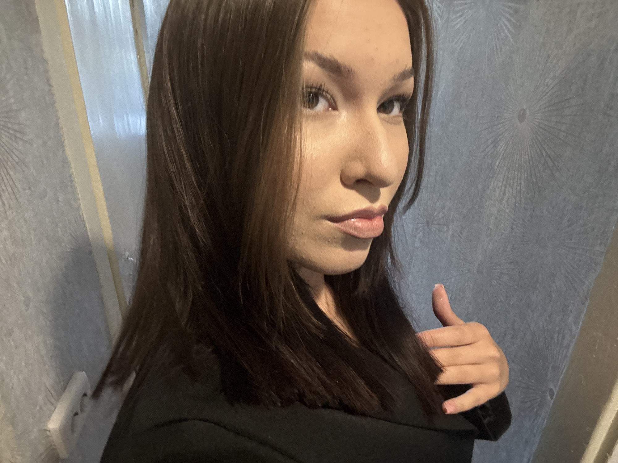 A young woman with dark brown hair is pictured from the chest up, looking towards the right of the frame with her head tilted. Her face is partially obscured by her hair, but her right eye, nose, and mouth are visible. She is wearing a black top. Her right hand is raised, giving a thumbs-up gesture. The background features a textured wall with a circular, sunburst-like pattern in a greyish-blue hue. To the left, a vertical cream-colored element, possibly a door frame or partition, is visible, along with a white electrical outlet on the wall. The lighting suggests an indoor setting, potentially a home in Inskoy, Russia.