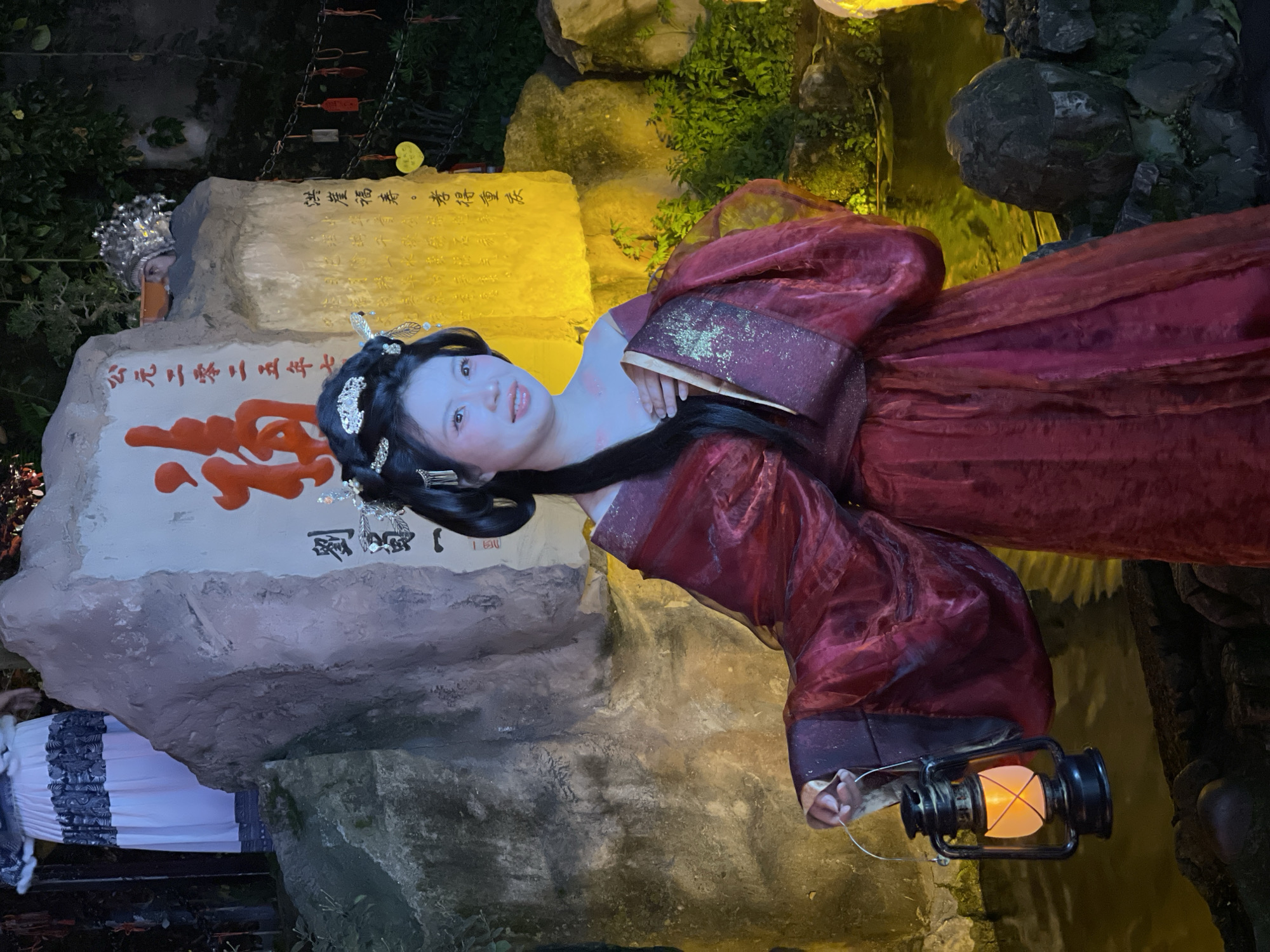 An East Asian woman, positioned centrally, smiles slightly while holding an illuminated black lantern in her right hand. She wears a dark red traditional Chinese-style garment with gold accents and an elaborate silver-toned headpiece in her dark hair. Her face is illuminated with a strong cool blue light, and her lips are light pink. Behind her is a large, irregularly shaped stone monument. The left face of the monument displays large red Chinese characters and smaller black text, including "July 2025 AD." The right face features black Chinese text, including "Hongya Fushou · How can we get Chongqing?", illuminated by a warm yellow light. Partially visible to the upper left is a person's lower torso wearing a white pleated skirt with a dark pattern. To the upper right, behind the monument, the top of a head adorned with a metallic silver headpiece is partially visible. The scene includes a dark body of water in the foreground and to the right, along with various rocks and green foliage. Small red and yellow decorative tags hang from strings near the right side of the monument. This nighttime setting is located in Yuzhong District, Chongqing, China, likely within the Hongyadong scenic area.