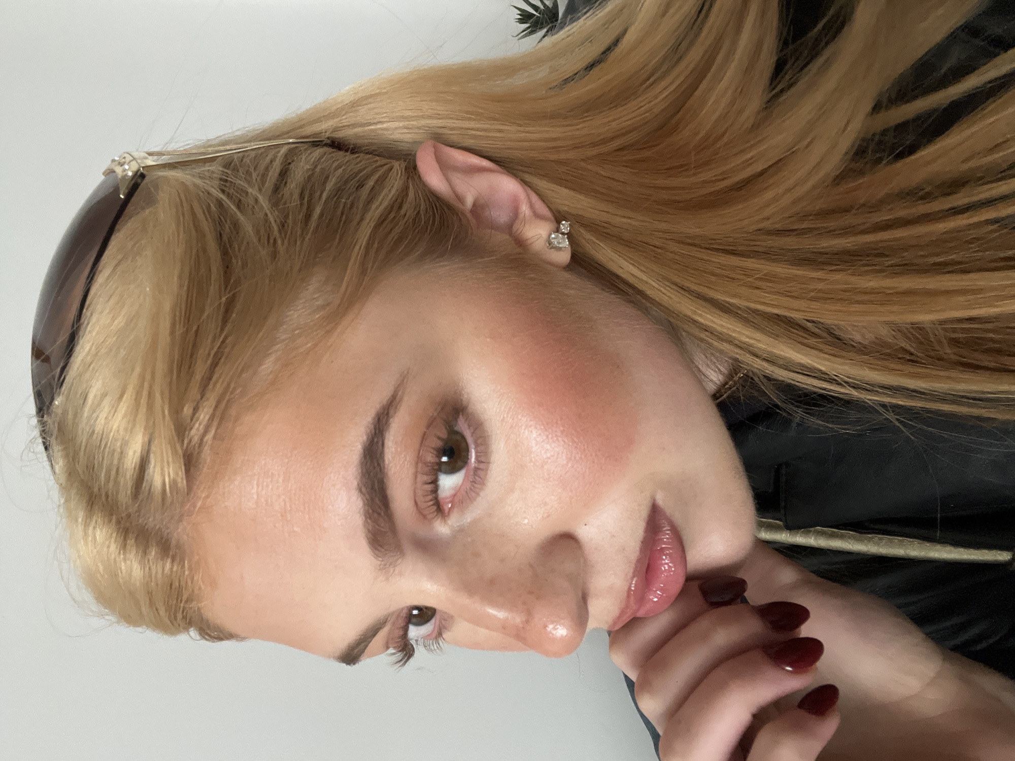 A close-up, angled portrait shows a fair-skinned woman with light brown eyes, freckles on her nose, and blonde hair. She has sunglasses perched on her head and is wearing an earring. Her nails are painted a dark red color, and she is wearing a black jacket. The background appears to be a white or light grey wall. There is no indication of the location being in Paris, France.