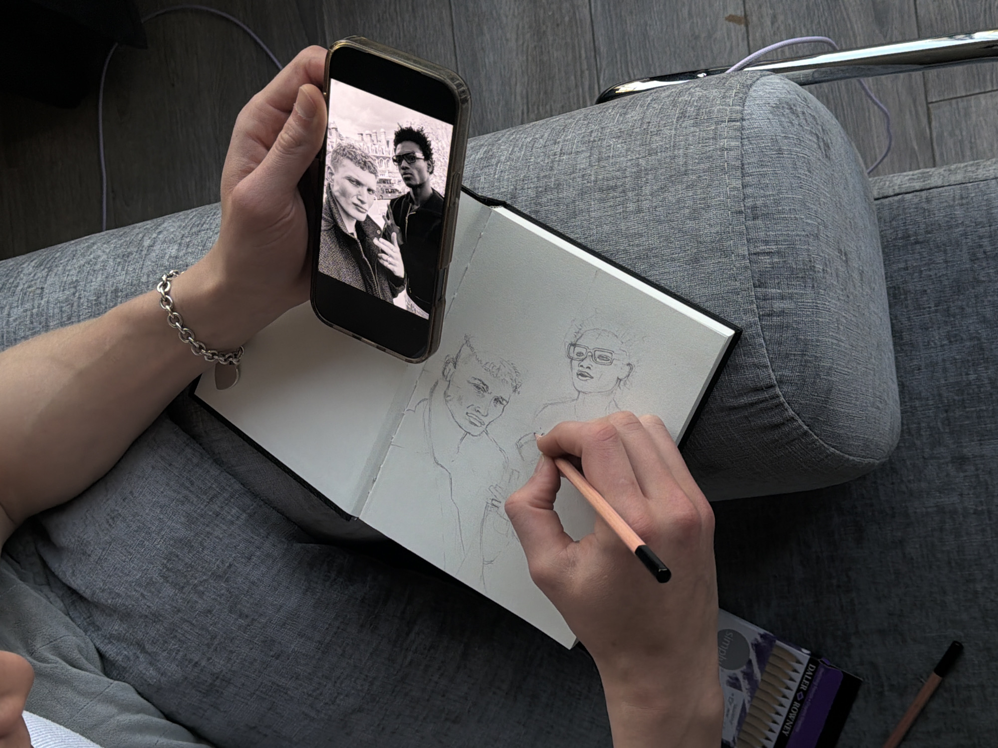 This overhead image depicts an individual engaged in drawing, likely in an indoor setting in London, United Kingdom. The artist's left hand holds a black smartphone, which displays a black-and-white photograph serving as a reference. The photo shows two men: one with short, light-colored textured hair looking subtly towards the viewer, and another with darker, curly hair and sunglasses, also facing the viewer. Buildings are faintly visible in the background of the phone's image.
The artist's right hand, adorned with a silver chain bracelet, holds a wooden pencil, actively sketching the likenesses of the two men onto an open sketchbook. The pencil drawing is in progress, showing the preliminary outlines of both figures' heads and shoulders. The person in the sketch on the right is depicted wearing glasses, mirroring the reference photo.
The scene is set against a backdrop of grey, textured fabric, suggesting a comfortable seating area such as a couch or cushion. Parts of a light wooden floor and a chrome bar with a purple cord are visible in the background. A box of "Daler Rowney Simply" pencils, indicating art supplies, rests near the artist's right hand. The image captures a moment of focused artistic activity.