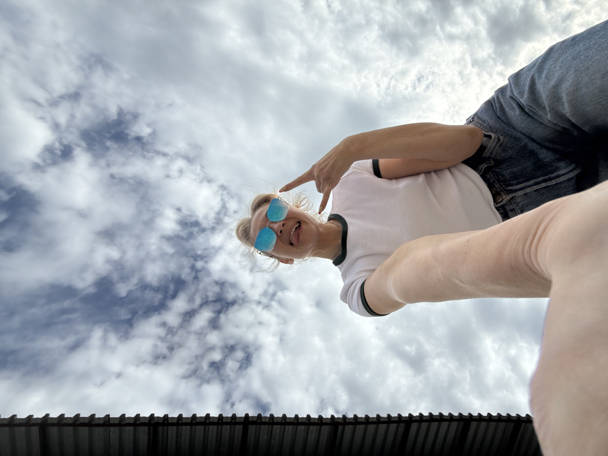 A low-angle, upward-facing shot depicts a person wearing blue mirrored sunglasses and a white t-shirt with black trim around the neckline and sleeves. The person is making a rock-and-roll hand gesture. Light blue jeans are partially visible. Above the person, a sky with varying cloud cover fills the frame. The bottom edge of the frame shows a dark, corrugated metal structure. While the location is supposedly in Chaikovsky, Russia, there are no discernible landmarks or indicators in the image confirming this location.