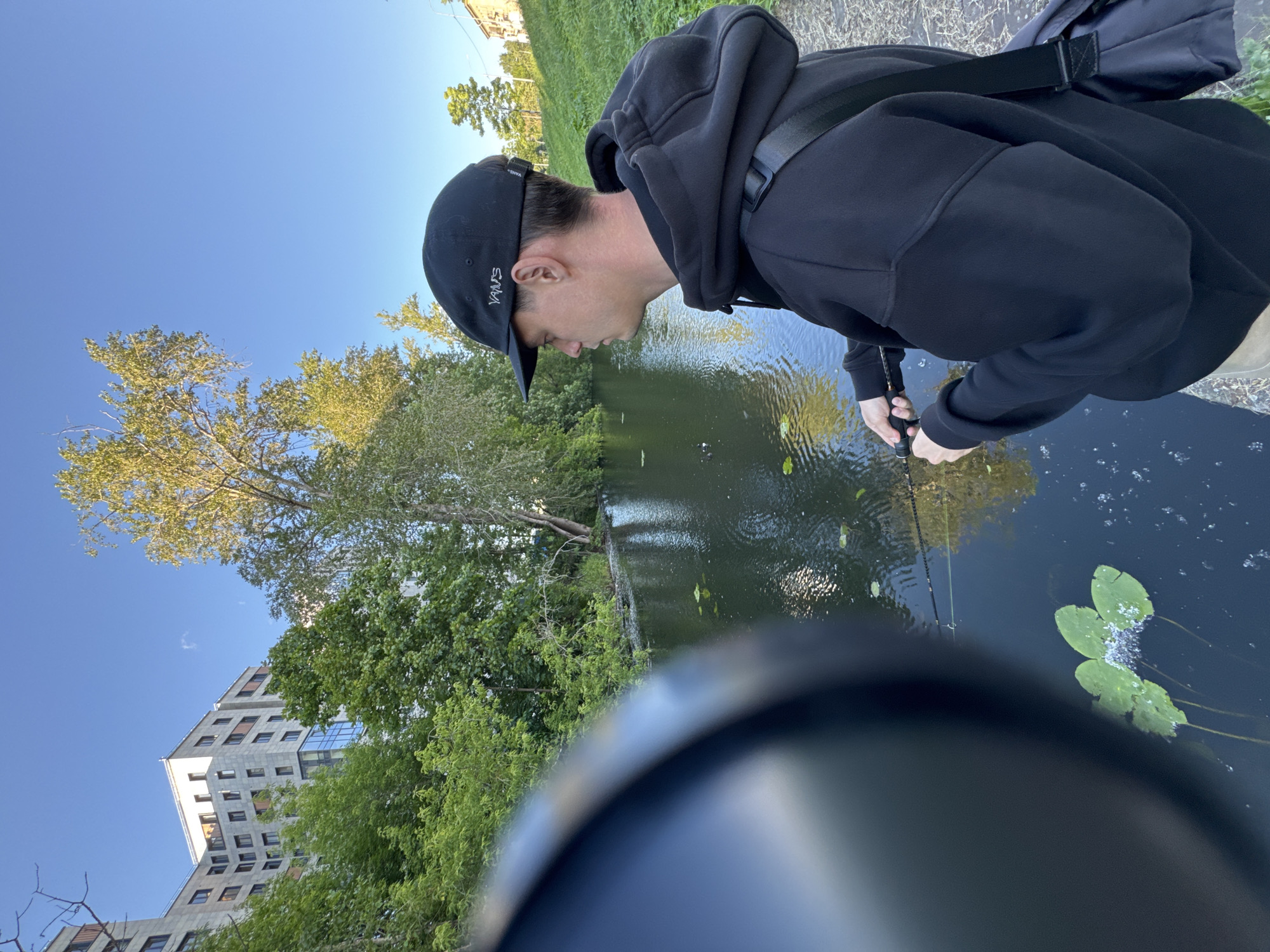 The image shows a person fishing in a canal in Saint Petersburg, Russia. The person, wearing a black hoodie and a black baseball cap, is focused on the water, holding a fishing rod. The canal is surrounded by lush green trees and a building with a modern architectural style, its facade a mix of gray stone and glass windows. The sky is clear and blue, suggesting a pleasant day, likely in the late afternoon or early evening given the warm lighting. The water is calm, with lily pads floating on the surface. The overall mood is one of peaceful activity in an urban natural setting. The visible text on the person's cap reads "VANS".