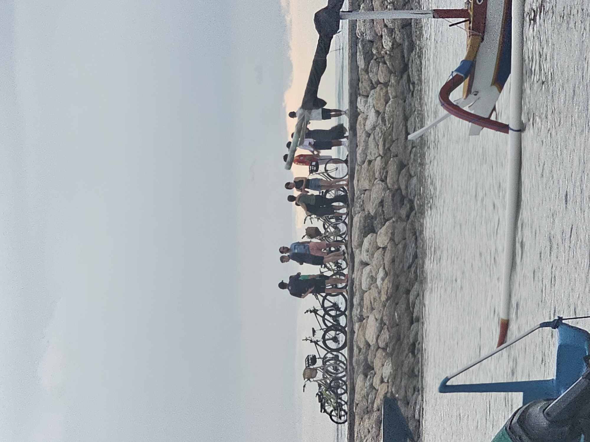 The image depicts a waterside scene, likely near the city of Denpasar, Indonesia. A group of about ten people are standing on a stone embankment at the water's edge. Many of them are accompanied by bicycles, which are lined up along the stone barrier. The individuals appear to be observing something in the distance. The weather seems overcast. Part of a boat is also in the frame.