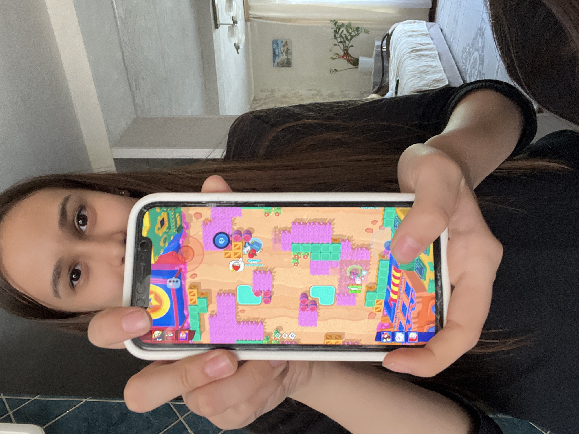 A woman is holding a smartphone displaying a game. She is indoors. Behind her, there is a glimpse of a room with furniture and wall decorations. The city and specific location in Chaikovsky, Russia cannot be determined from the image.