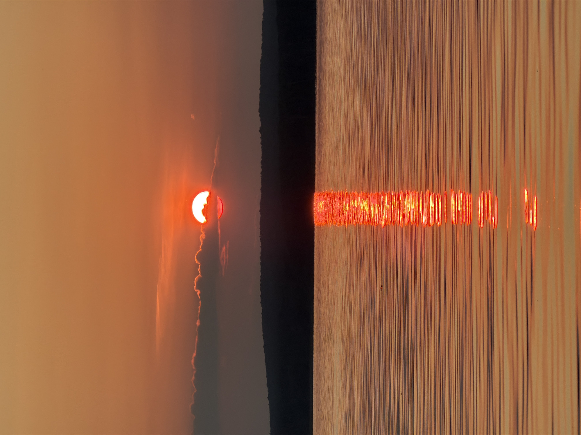 A sunset is captured over a calm body of water, likely the Kama River, near the city of Chaikovsky, Russia. The sun, a bright orange orb, is partially obscured by a thin layer of clouds and sits just above the dark silhouette of a distant tree line. Its reflection creates a shimmering vertical path of light on the water's surface, broken by gentle ripples. The sky transitions from a soft orange near the horizon to a warmer tone higher up. The water itself reflects the warm hues of the sky and sun, creating a tranquil and atmospheric scene. No people are visible in the image.