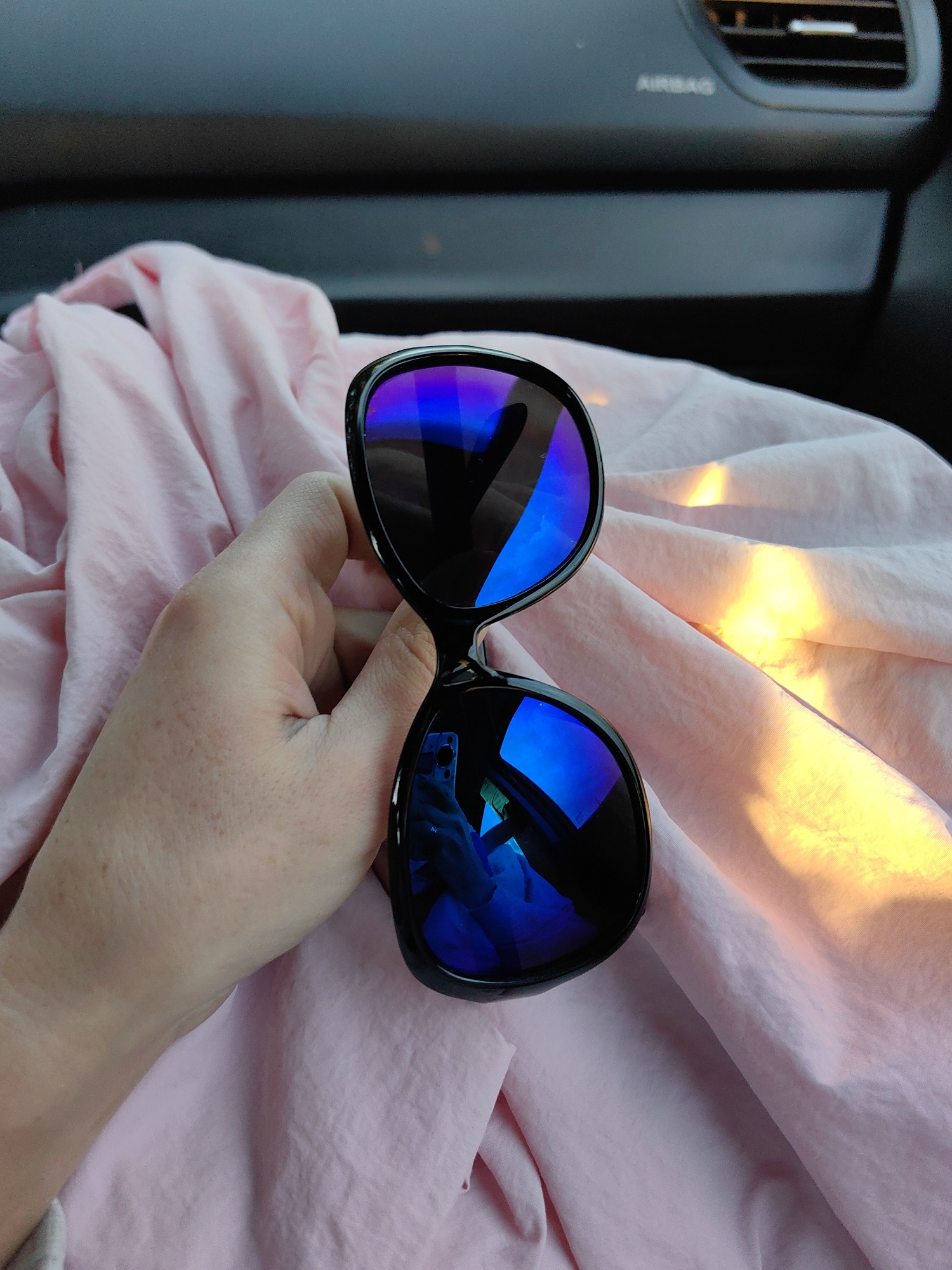 A person's hand holds a pair of black-framed sunglasses with reflective blue lenses. The sunglasses are resting on a crumpled piece of light pink fabric, which appears to be a scarf or piece of clothing. In the background, a dark gray car dashboard is visible, with the word "AIRBAG" printed in white. The scene is illuminated by warm, golden sunlight, casting bright highlights on the fabric. The overall impression is an indoor or automotive setting, with the focus on the sunglasses and their reflection. The location is not discernible from the image itself.