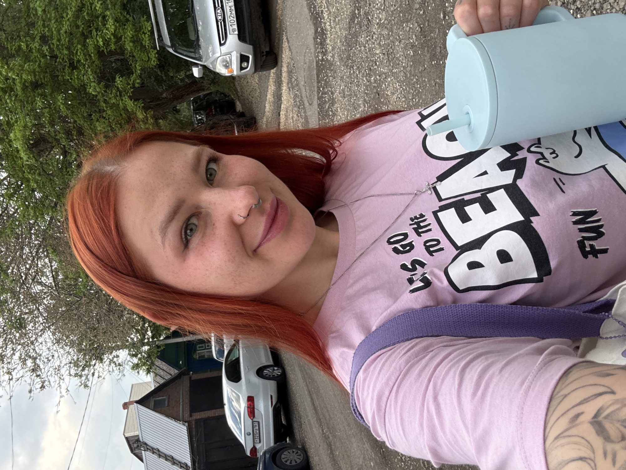 A woman with red hair and a nose ring is taking a selfie outdoors. She is holding a light blue drinking cup with a straw. She is wearing a pink t-shirt with a cartoon design and text that reads "Let's go to the beach fun". The background includes parked cars, trees, and a building. Based on the license plates of the cars present, it is likely that this image was taken in Rostov-on-Don, Russia.