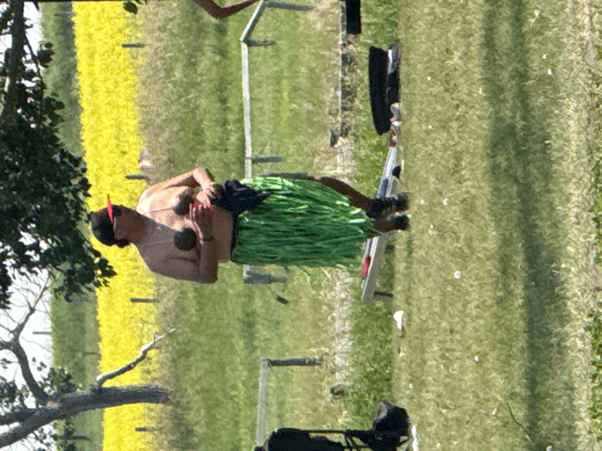 A person wearing a black baseball cap with a red brim and sunglasses is standing in a grassy area. They are shirtless and wearing a green grass skirt. They are holding two round objects in their hands. In the background, there is a field of yellow flowers and trees. The ground is covered in green grass with scattered white flowers and some fallen leaves. There are also some wooden posts and fences visible in the background. The image appears to be taken outdoors in a rural or park-like setting. The location is not specified in the image.