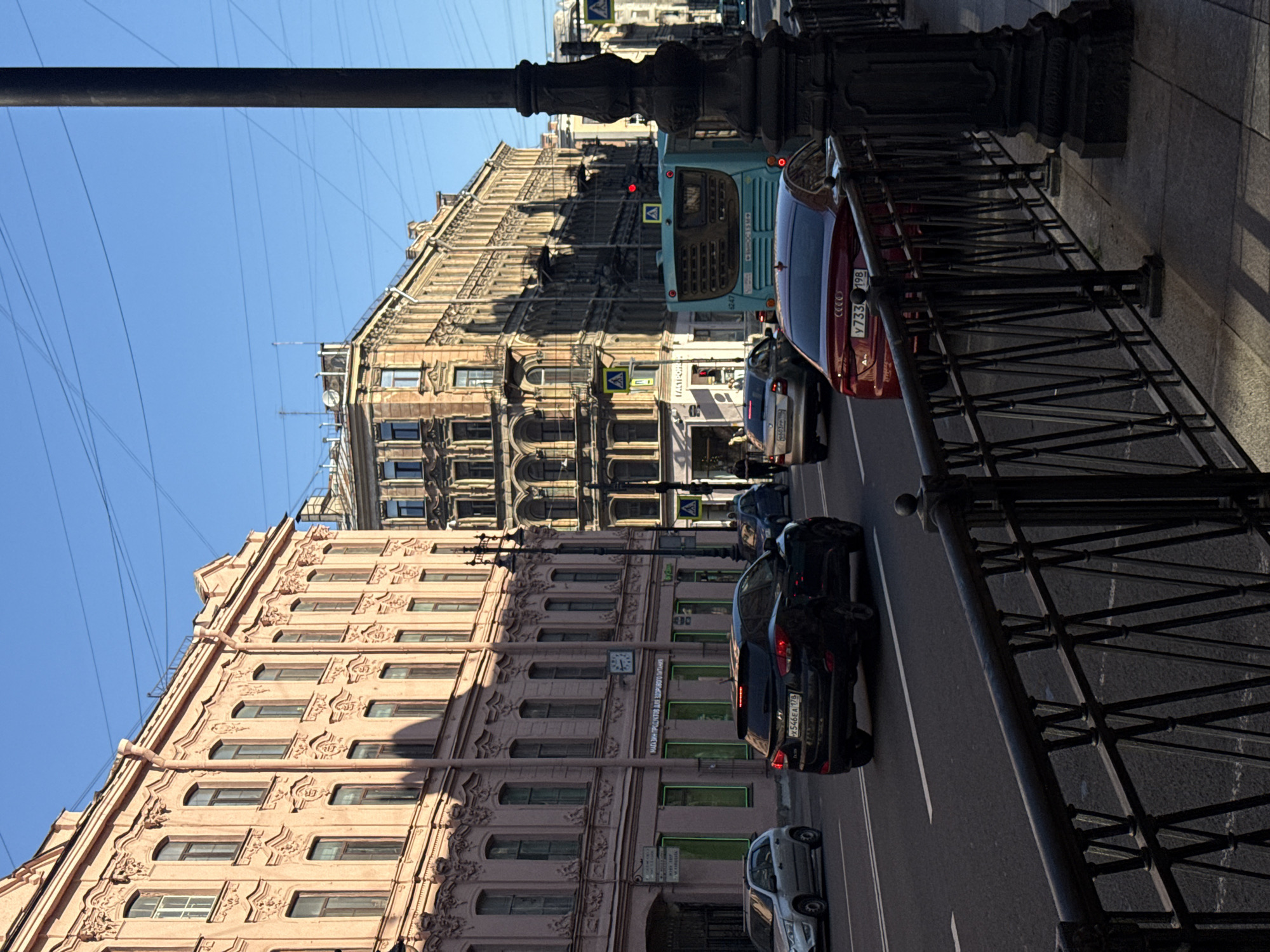 In the city of Saint Petersburg, Russia, on a clear day, this photograph captures a street scene. The perspective is from sidewalk level, looking towards a city street with several buildings and vehicles.
On the left, a pale pink building with ornate architectural details and numerous windows lines the street. Across the street, there are other buildings with a different architectural style, appearing older and more weathered. A light blue bus is stopped in traffic. Several cars are visible on the road, including a black SUV, a red sedan, and a silver hatchback. Pedestrians are present on the sidewalk, including a person walking on the right side of the frame.
The environment is urban, with paved roads, sidewalks, and streetlights. The sky is clear and blue, indicating fair weather. The overall impression is one of a typical day in a historic city.
