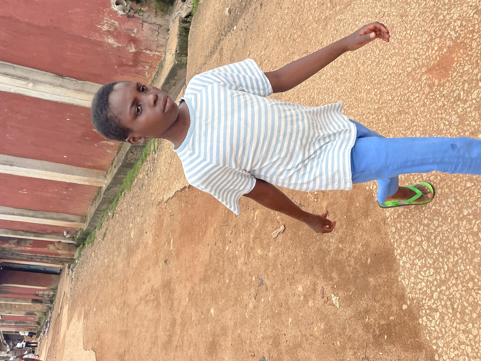 A young person, appearing to be of African descent and pre-pubescent, walks down a dirt road in a residential area of Jos, Nigeria. The person is wearing a blue and white striped t-shirt and solid blue pants, along with bright green flip-flops. They are looking to their right, with a neutral expression. The road is unpaved, with patches of gravel and reddish-brown dirt. To the right of the road, there is a weathered, red-painted building with concrete accents and some signs of decay. The background shows more of the building and hints at other structures and vegetation, suggesting a typical urban environment in Jos. The lighting indicates it is daytime, with diffused light suggesting either overcast conditions or late afternoon.