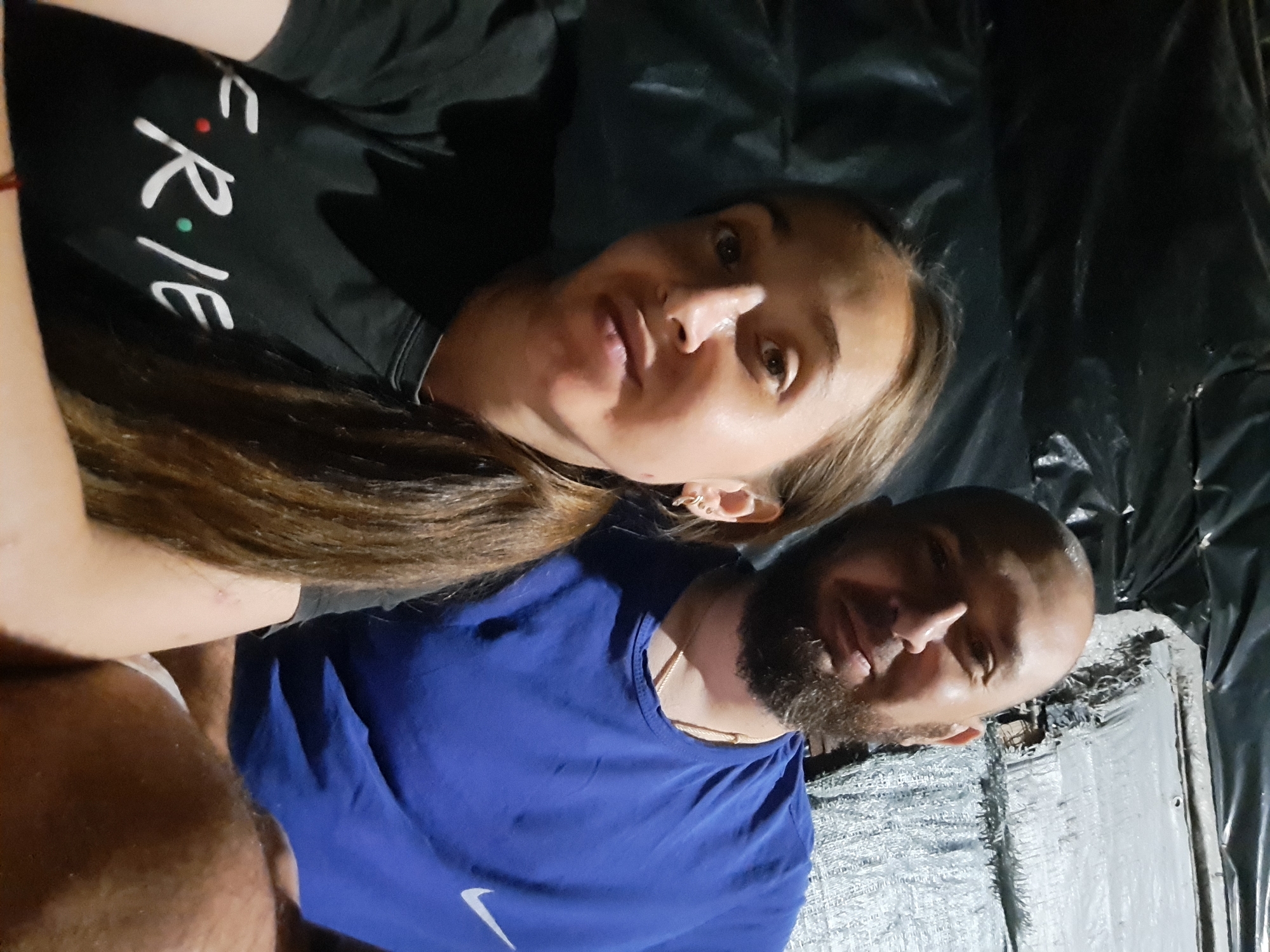 The image shows a selfie of a man and a woman taken indoors. The man, who is bald and has a beard, is wearing a blue t-shirt with a Nike logo. The woman, with long brown hair, is wearing a black t-shirt with the word "FRIENDS" written on it. They appear to be in close proximity. The background is dark and appears to be a rough, unfinished wall or structure, possibly a shelter or makeshift accommodation, with black plastic sheeting visible. The lighting is dim. The location is identified as Vypasne, Ukraine.