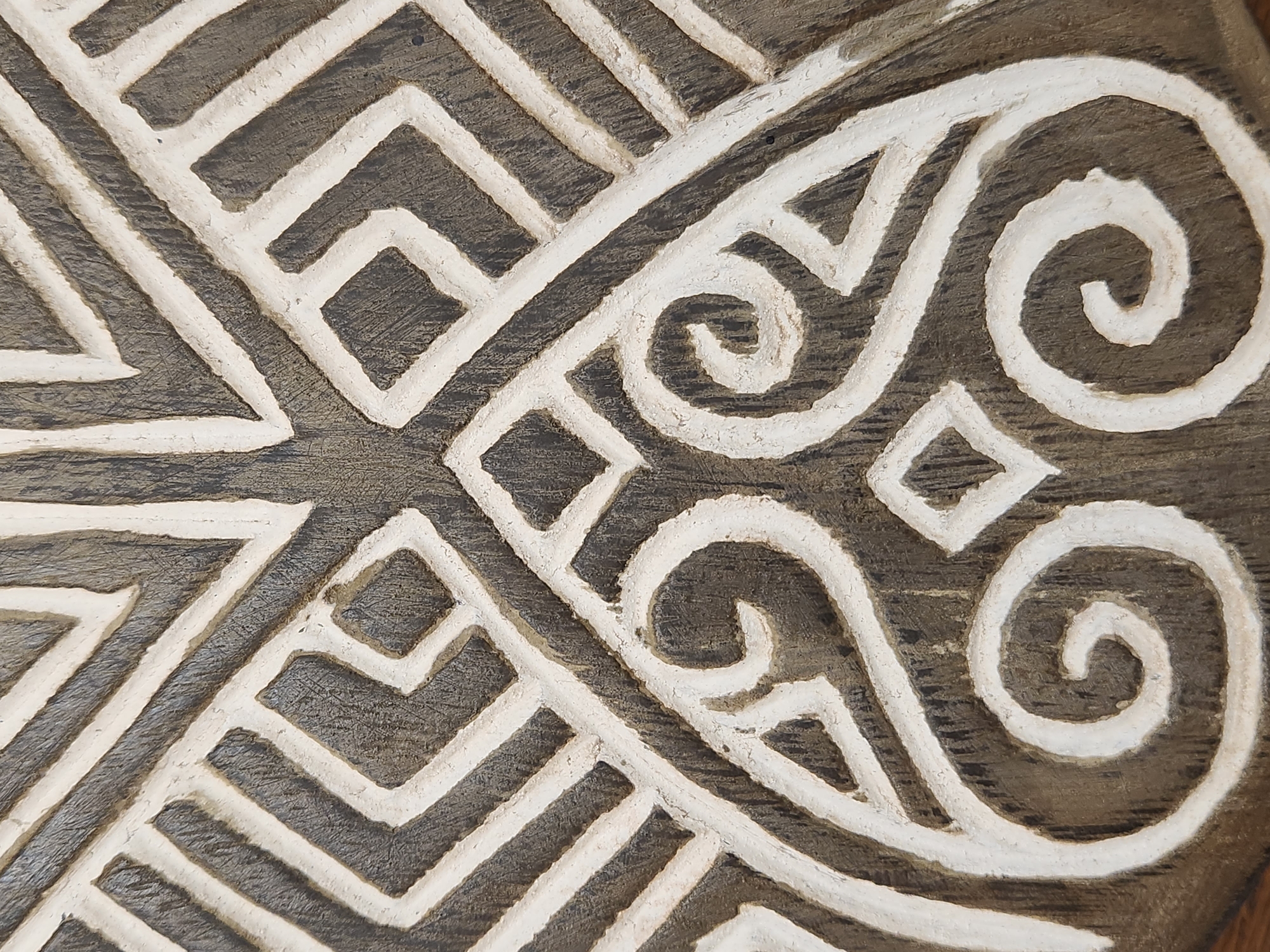 The image shows a close-up view of a carved wooden object with a white, geometric pattern. The wood appears to be dark brown and has a visible grain. The carving is raised and the white paint is applied to the raised surfaces, creating a strong contrast. The pattern consists of interlocking lines, diamonds, and swirling shapes. The texture of the wood and the carving is evident, with some imperfections and rough edges visible.
There is no discernible information in the image to suggest a specific location in Maitland, United States of America. The image focuses solely on the texture and pattern of the carved object.