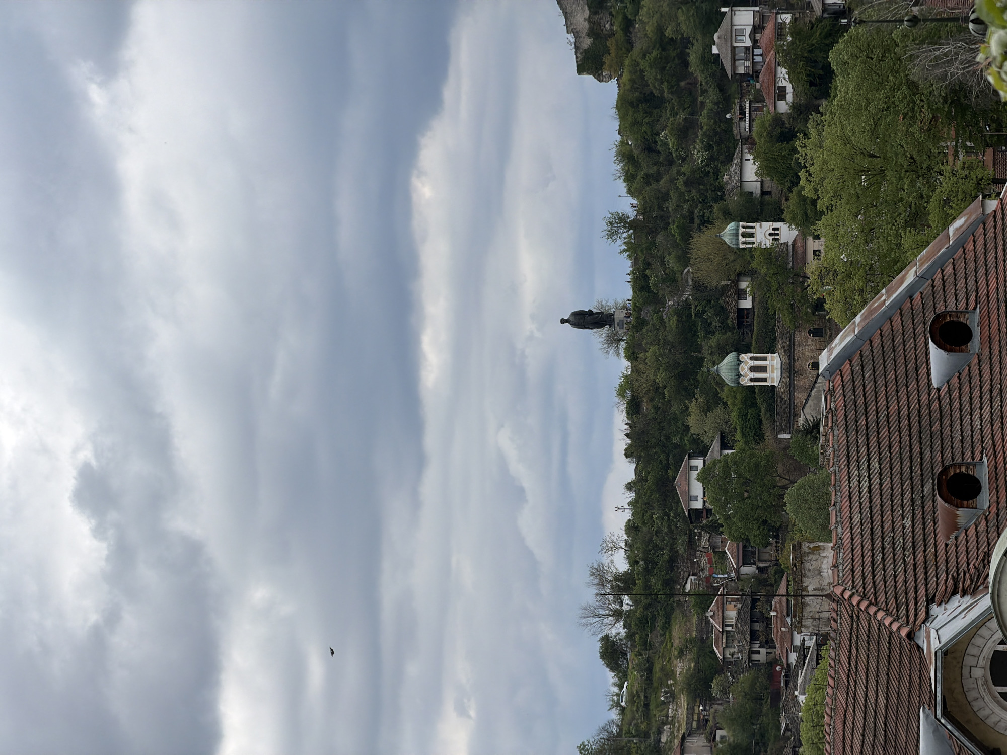 The image presents a view of the old town of Lovech, Bulgaria, under a cloudy sky. A statue stands atop a verdant hillside, overlooking the town. The foreground features rooftops with red tiles and chimneys. Traditional houses are interspersed with trees on the hillside.