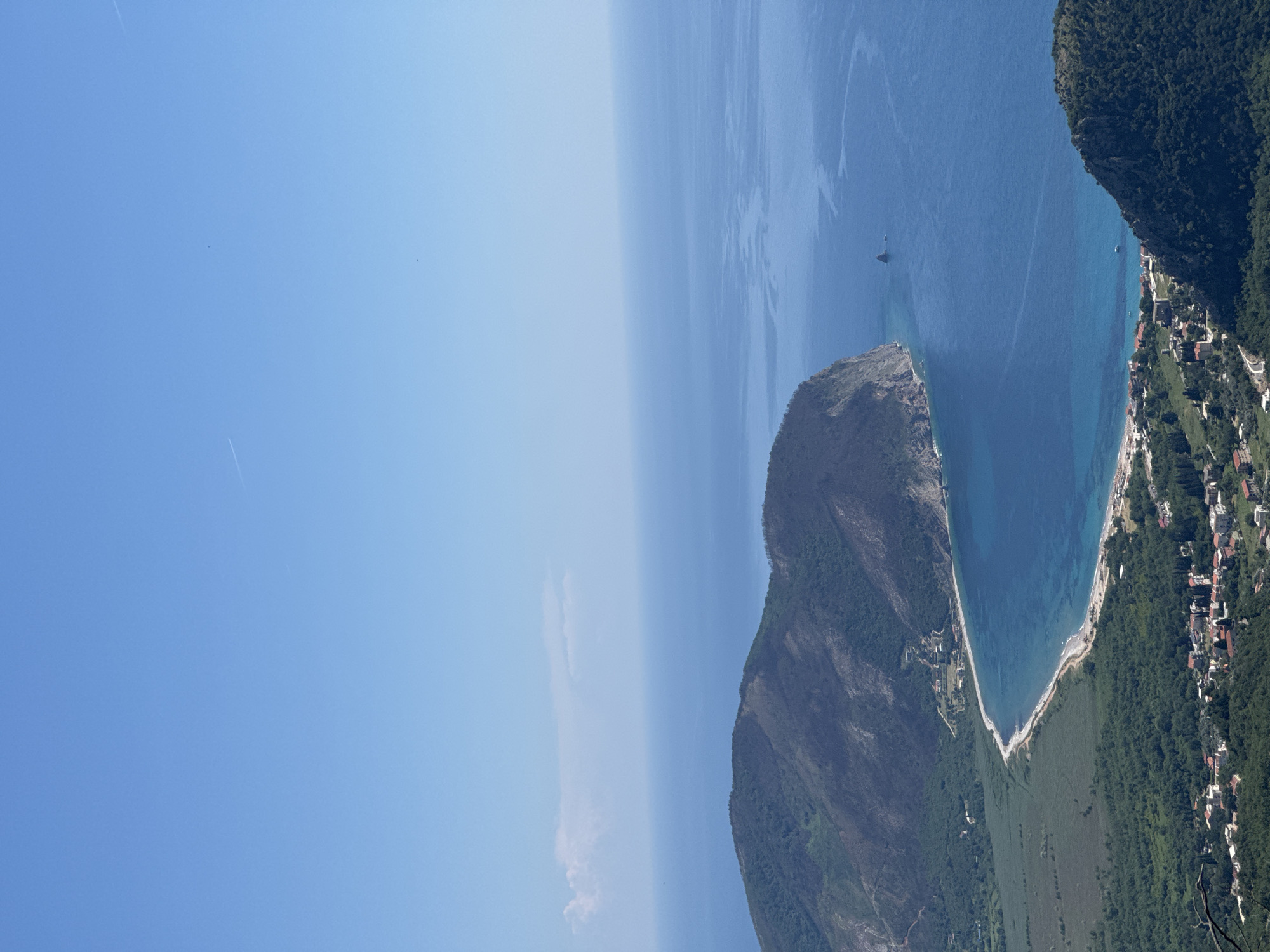 This aerial shot, taken in Novoselje, Montenegro, shows a coastal scene. On the right, a village nestled on the hillside edges a strip of beach lining a bay. The blue water of the bay leads to the open sea. To the left of the bay, a rocky peninsula covered with sparse vegetation juts into the water. The sky, predominantly clear blue, occupies the left side of the image. A single sailboat is seen in the bay.
