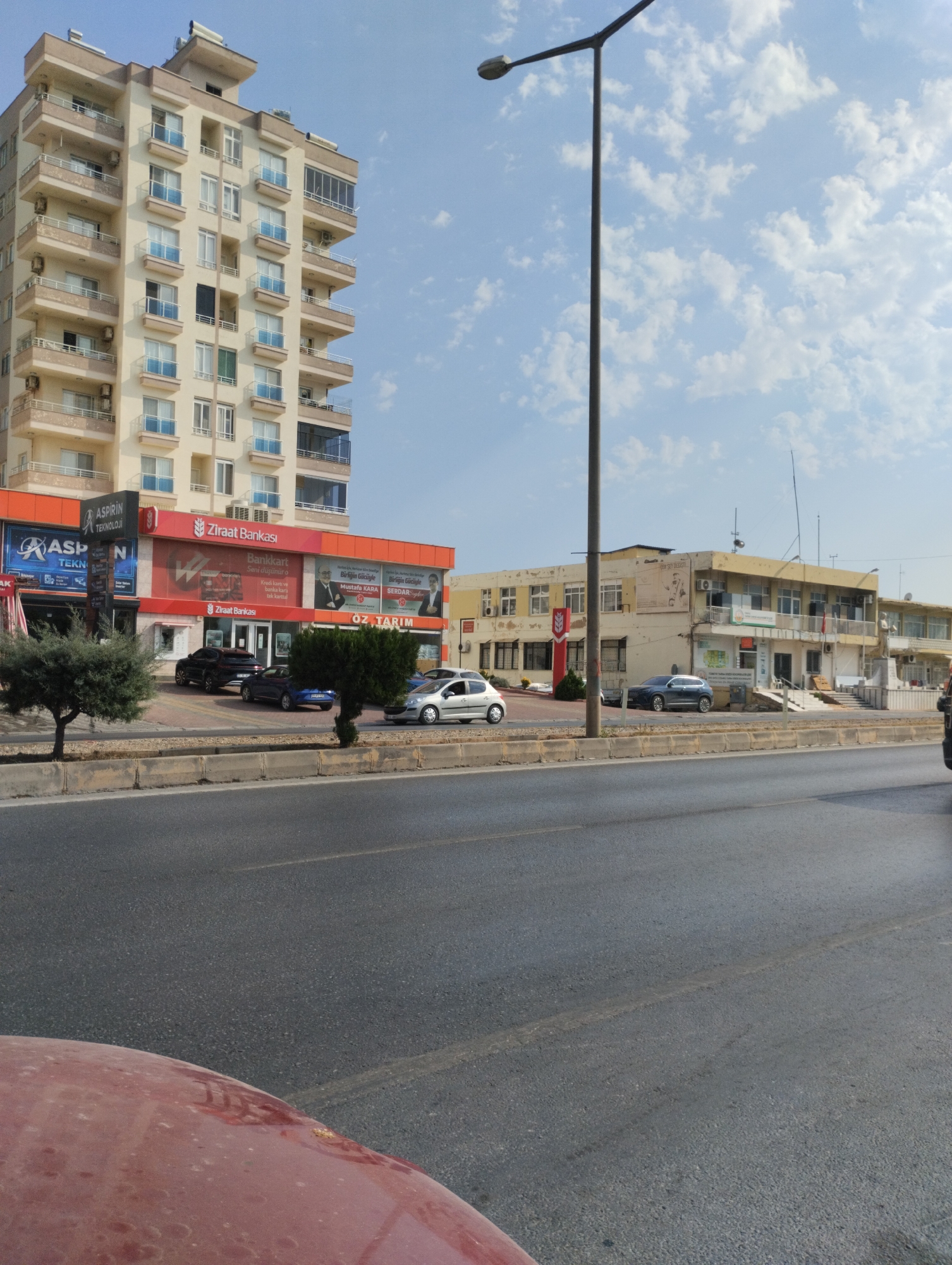 In the city of Erdemli, Turkey, on a clear day with scattered clouds, a street scene unfolds. A tall, multi-story building with beige and cream-colored walls dominates the left side of the frame. It features numerous balconies with light blue railings and various windows. Street-level businesses are visible, including "Ziraat Bankası" with its red signage, and "ASPİRİN TEKNOLOJİ." A street lamp with a dark pole and a light fixture stands tall in the center. To the right of the tall building is a lower, yellow building with multiple windows and some signs, one appearing to be a statue at its entrance. Several cars are parked in front of the businesses and on the street, including a silver sedan, a dark sedan, and a blue sedan. Green trees and bushes are planted along the sidewalk. The foreground shows a portion of a reddish vehicle and a gray asphalt road with visible tire marks and lane lines. The overall impression is a typical urban streetscape with commercial buildings and vehicular traffic.