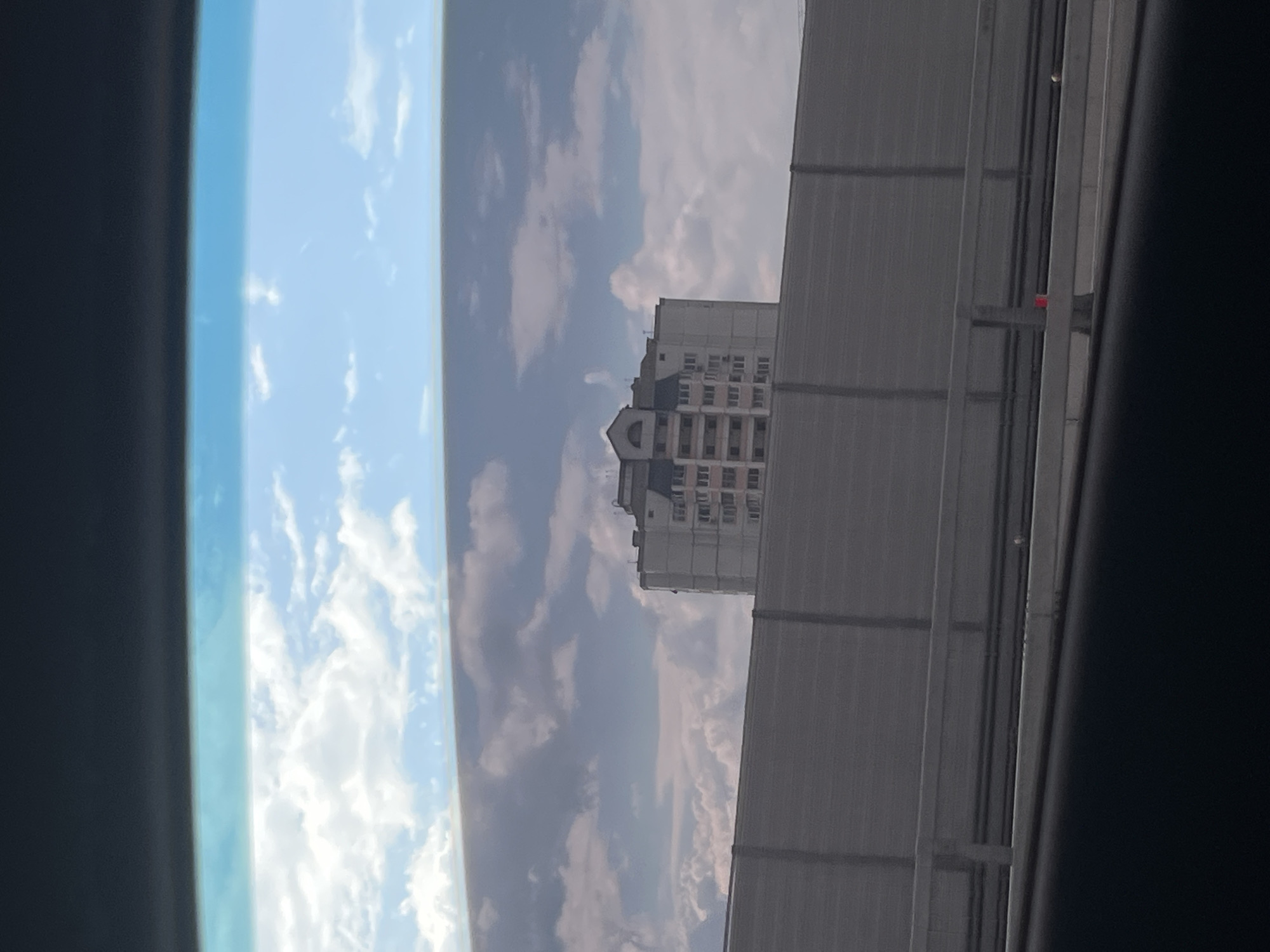 The image, taken from inside a vehicle, shows a multi-story apartment building in Moscow, Russia. The building, predominantly light-colored with darker accents around the windows and roofline, features numerous windows arranged in a grid pattern. A distinctive pitched roof with dormer windows is visible at the top. In the foreground, a grey sound barrier runs horizontally across the frame, obscuring the lower portion of the building and the ground. Above the sound barrier and the building, a bright blue sky is dotted with white, fluffy clouds and darker, heavier clouds. The overall scene suggests an urban environment, possibly viewed from a highway or elevated road.
