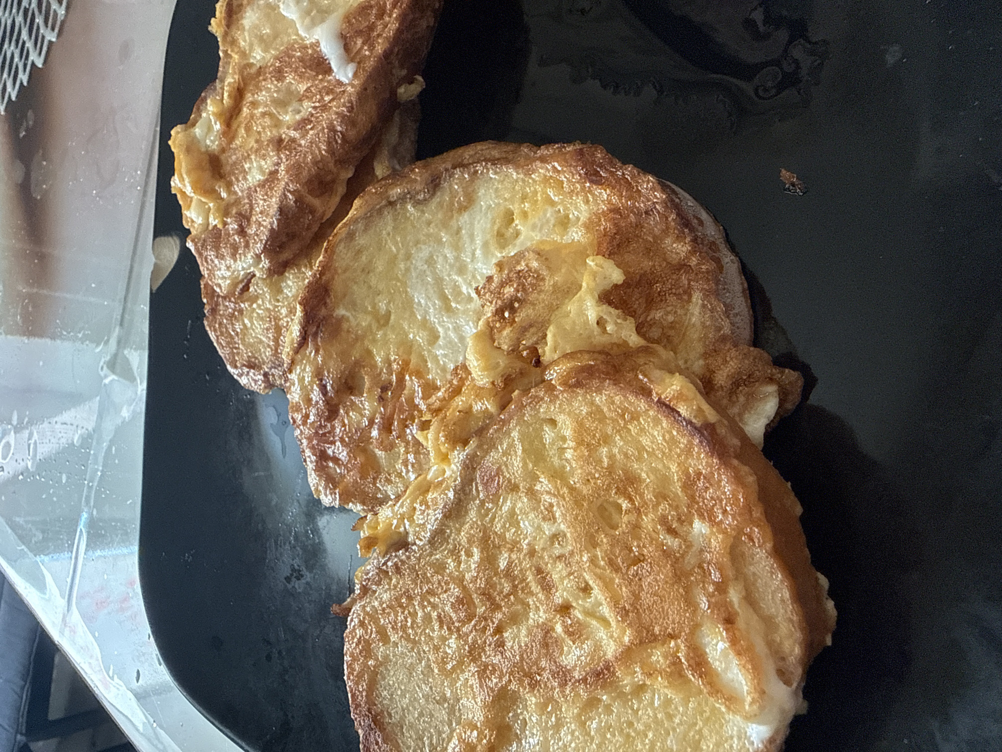 A black plate with three slices of golden-brown French toast sits on a reflective surface. The French toast is slightly uneven in color, indicating it has been fried. The surface the plate rests on is wet and reflects the French toast and the plate. The scene appears to be indoors, with a blurry background of a kitchen counter. The overall impression is of a simple, homemade breakfast. There is no information about the location in Simferopol, Ukraine.