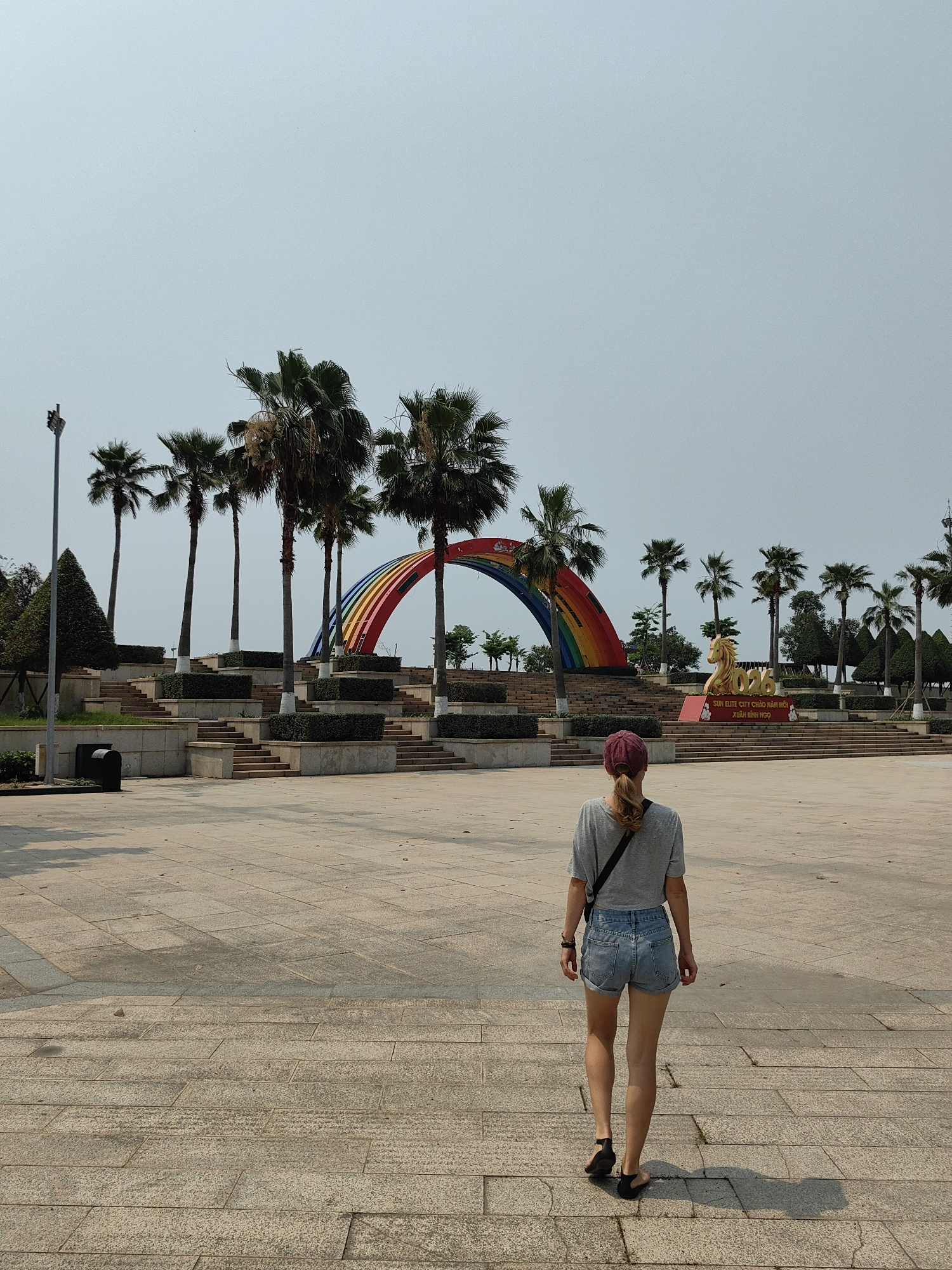 The image shows a woman from behind, walking away from the viewer on a large, paved area in Bai Chay Ward, Vietnam. She is wearing a grey t-shirt, denim shorts, a maroon cap, and black sandals. A black strap of a bag crosses her back. In the background, there is a multi-tiered structure with palm trees and shrubs. A large, colorful rainbow arch is positioned behind the trees. Further back, a golden horse sculpture and the number "2026" are visible on a raised platform. The sky is overcast. There is a trash receptacle on the left side of the frame.