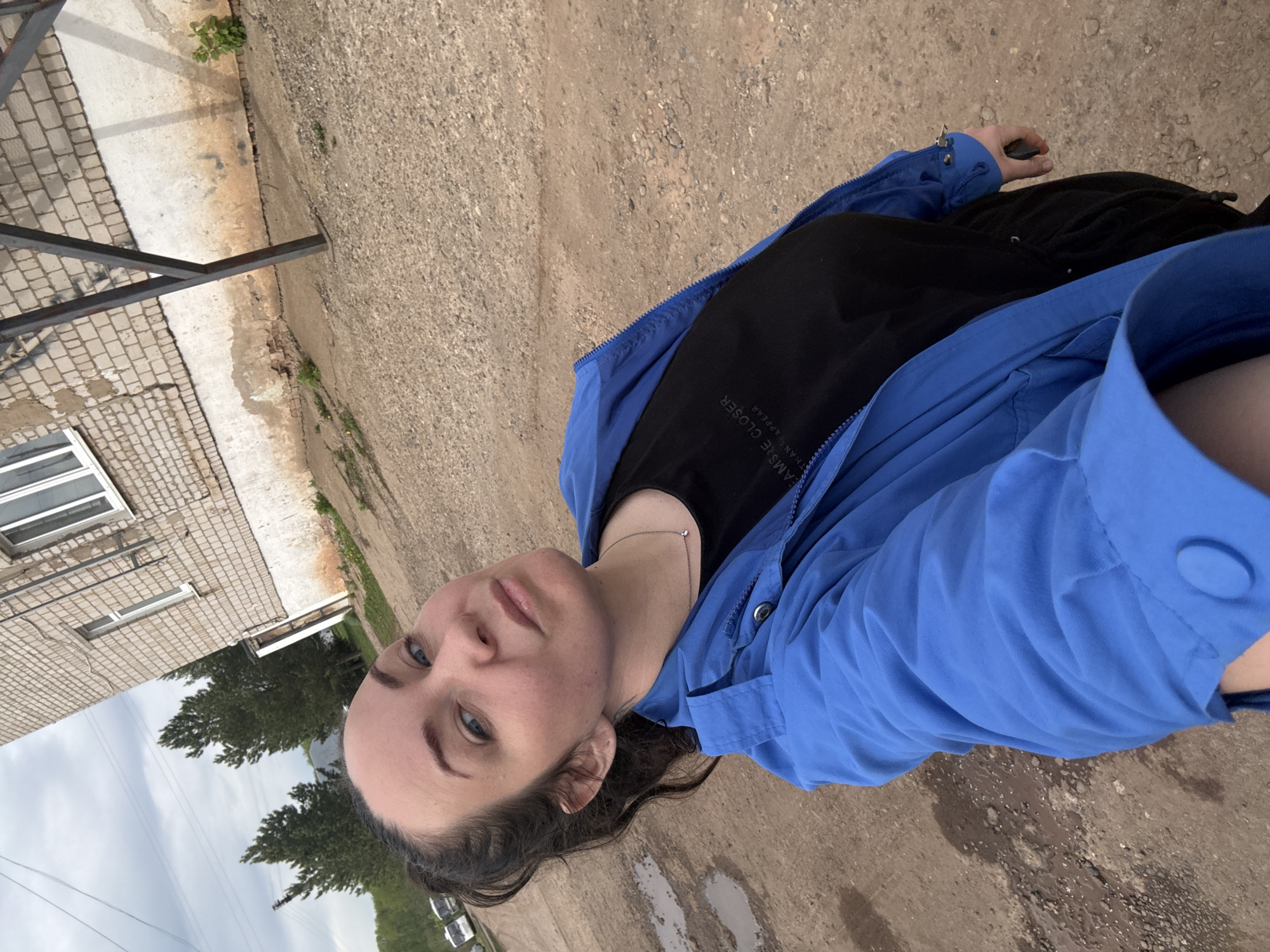 The image shows a woman outdoors in Фоки, Russia. She is wearing a blue jacket over a black shirt, with black pants. The ground appears to be dirt with some puddles. A brick building is visible in the background along with trees and a cloudy sky.