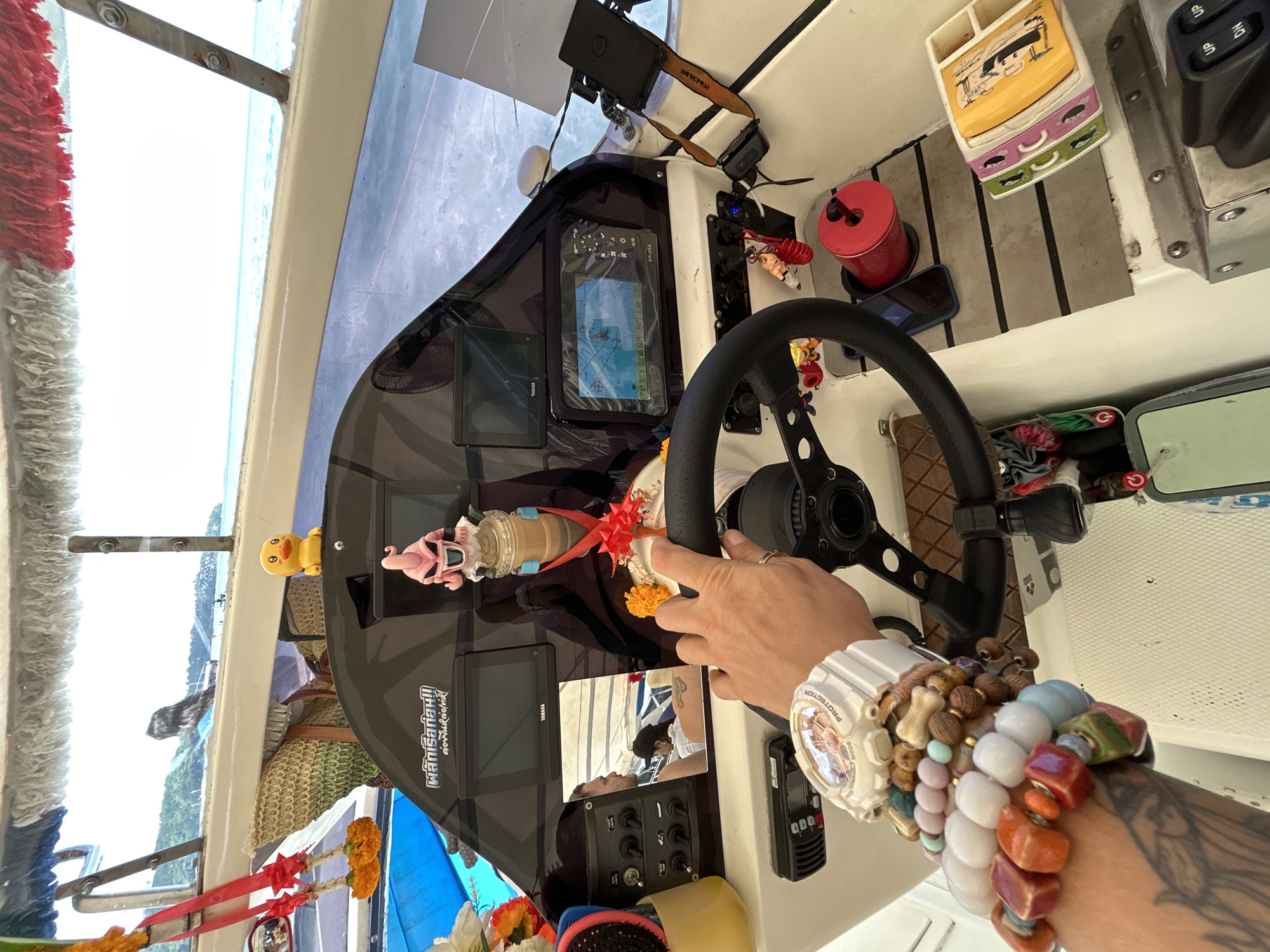 The image depicts the interior of a boat, likely a speed boat, on the water near Ko, Thailand. The foreground is dominated by a person's arm, adorned with numerous colorful bracelets and a white watch, resting on the steering wheel. The arm is tattooed with what appears to be floral designs.
The boat's dashboard is visible, featuring several electronic displays, including a GPS unit showing navigation data. A small, pink figurine resembling Majin Buu from Dragon Ball Z is placed on the dashboard, alongside a yellow duck figurine. Two red ribbons with orange flowers are tied to the boat's structure. A photograph of two individuals is also visible on the dashboard.
Other objects on the boat include a red cylindrical container, a dark-colored smartphone, a black radio or communication device, and a compartmentalized tray with what looks like small figures or images. In the background, the calm blue sea and a lush green island coastline are visible, characteristic of the tropical scenery in Thailand. The overall impression is a snapshot of a leisurely boat trip in a scenic, tropical setting.