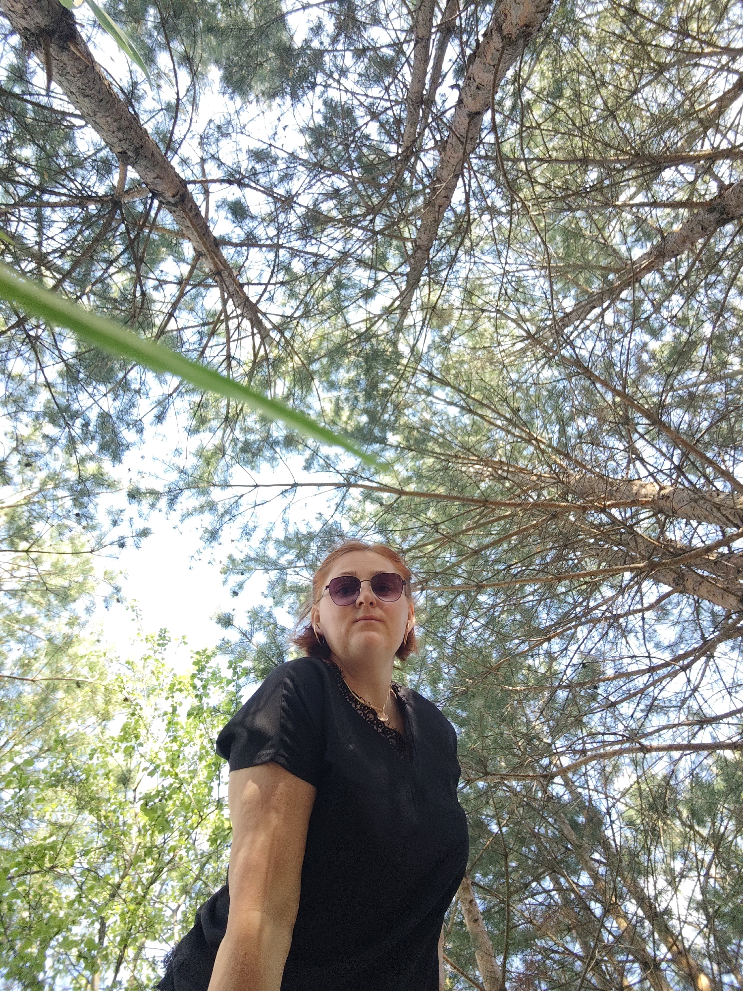 The image shows a woman with red hair and sunglasses wearing a black shirt. She is standing outside, looking up at trees with many branches. The location is Марковский, Russia. The sky is visible through the branches.