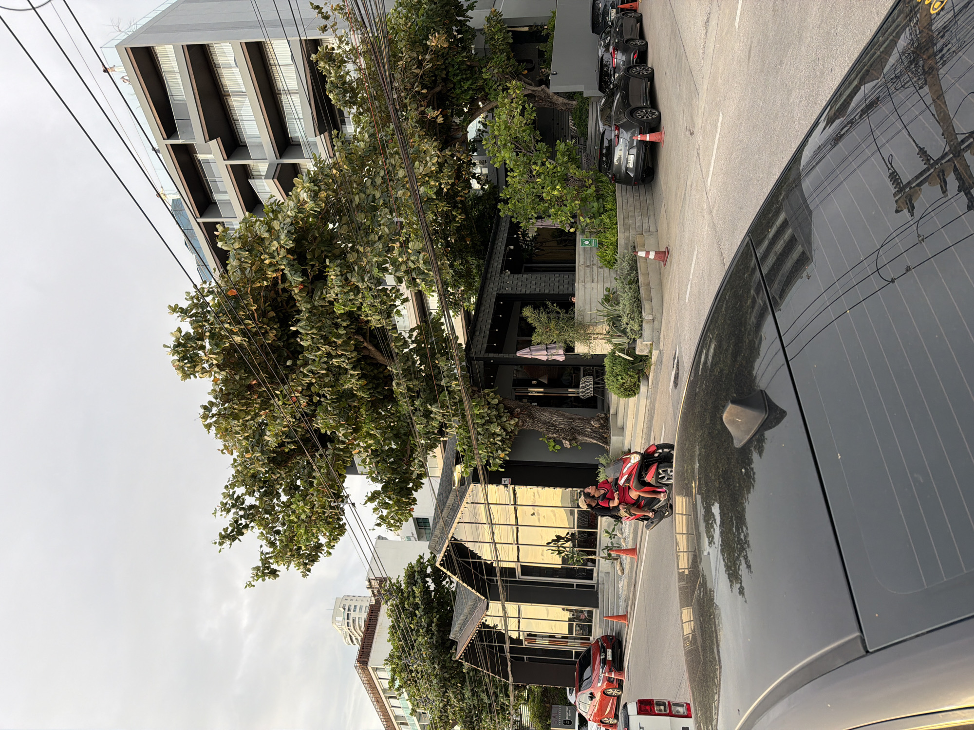 The image shows a street scene in Pattaya City, Thailand. A multi-story building is on the left, with trees lining the sidewalk. A dark-colored car, is positioned in the right foreground, obscuring part of the view. Several other vehicles are parked along the curb. Two people are on a red motorbike. The reflections on the car windows show overhead power lines. The sky is overcast.