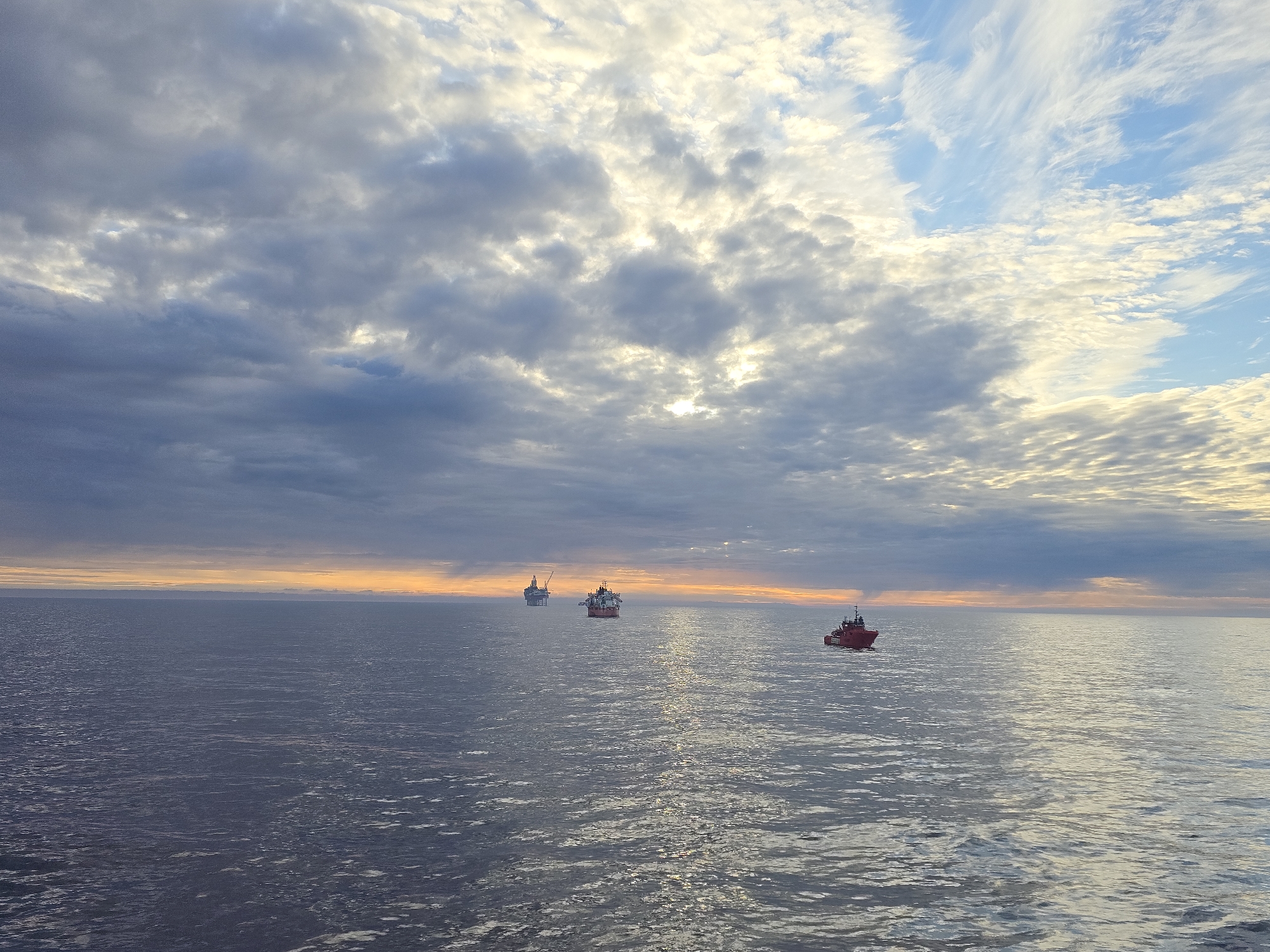 The image depicts a seascape under a sky filled with varying cloud formations, including cirrus and cumulonimbus clouds. The horizon line is slightly blurred due to atmospheric haze. Three vessels are visible on the water. One is a dark structure, likely an offshore platform, in the far distance. Another, smaller vessel is closer in view, and a red boat is positioned on the right side of the frame. The water surface is rippled, reflecting the light from the sky. The location is Unknown, Unknown.