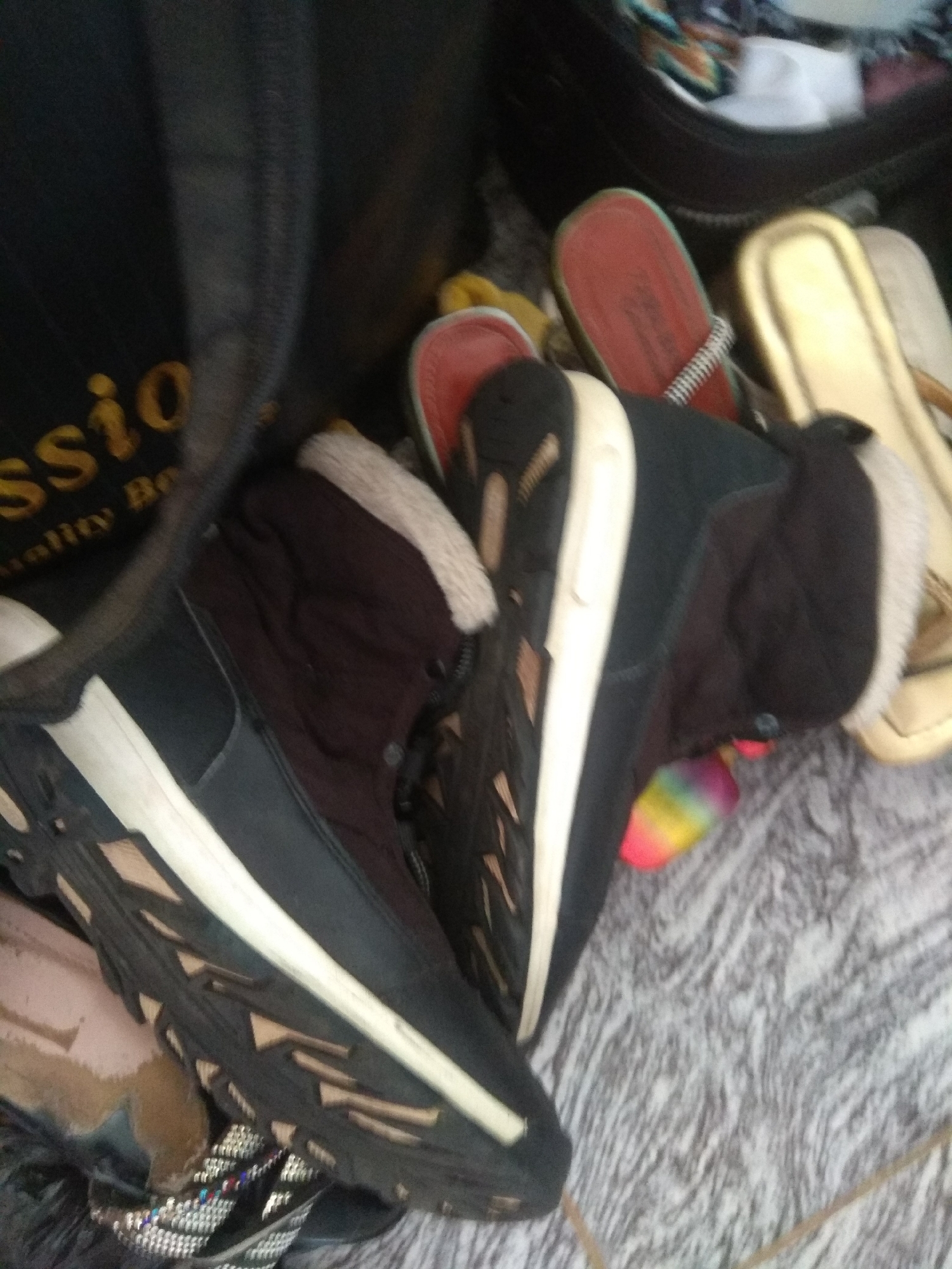 The image displays a close-up view of several pairs of footwear, appearing to be inside a bag or container, possibly a duffel bag given the visible fabric texture and stitching. In the foreground, a black and white sneaker-like shoe is prominently featured, with a thick, textured sole and laces. To its right, a dark boot-like shoe with a white furry lining is visible, partially obscured by the sneaker. Further back and to the right, two pairs of sandals are visible. One is a bright red sandal with a white and black patterned strap, and the other is a golden-colored sandal. The background consists of the dark fabric of the bag and a patterned surface beneath the footwear, resembling a carpet or mat. The scene appears to be indoors. There is no discernible information within the image to indicate a specific location in Jalingo, Nigeria, or any details about people, actions, or interactions.