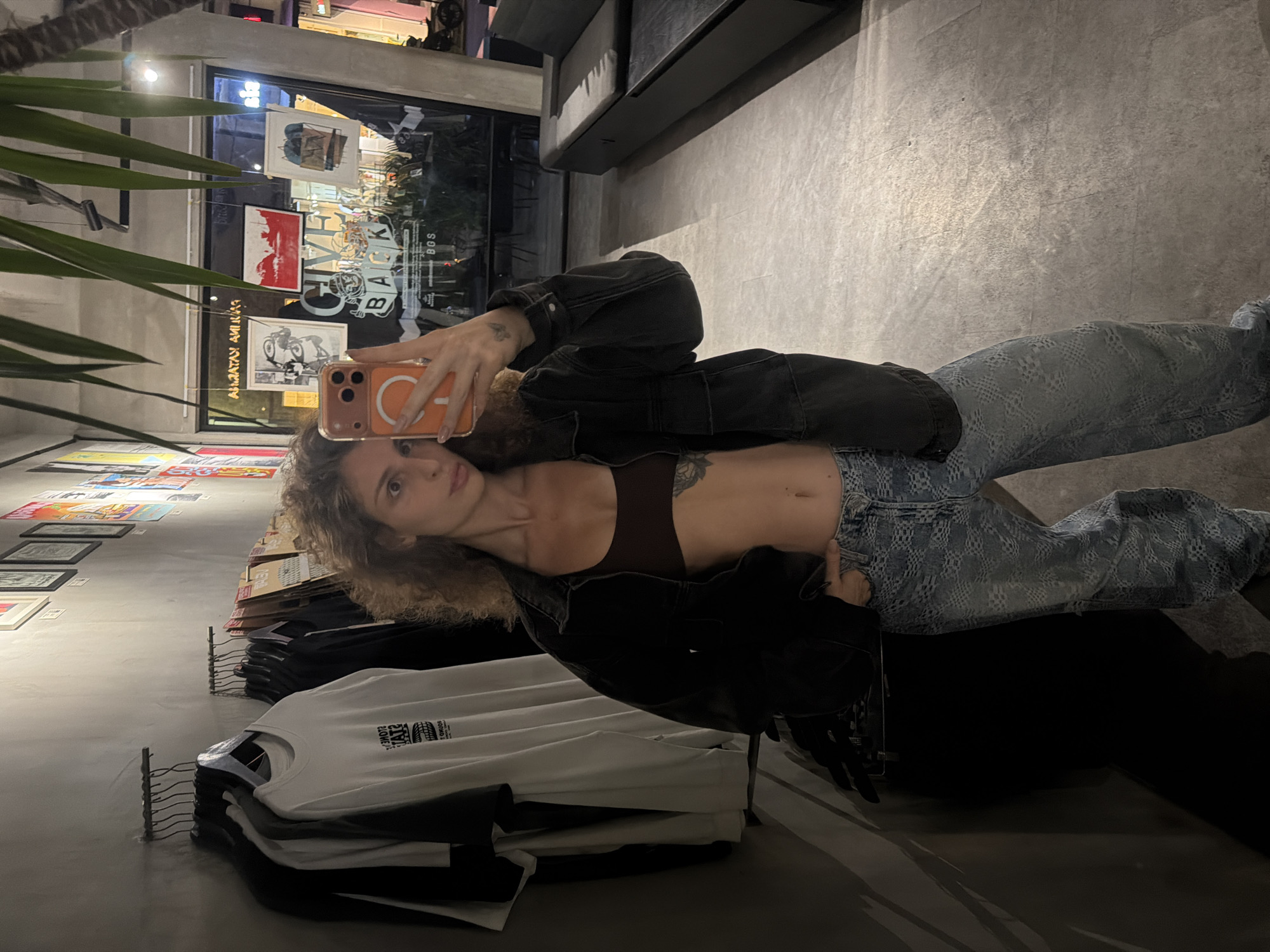 A person is taking a mirror selfie in a store. The person is wearing a dark jacket over a brown top, and blue jeans with a textured pattern. Their hair is curly and light brown. They are holding up a smartphone with an orange case. In the background, there are racks of folded t-shirts, some displayed on the wall, and posters. The setting appears to be a retail space, possibly a clothing store, with a modern, industrial aesthetic, characterized by concrete floors and walls. The lighting is dim, suggesting it might be evening or indoors. Visible text includes "CIVIC BACK BOS" on a poster in the background, and text on the t-shirts which is difficult to read. The location context is North Kuta, Indonesia. The overall mood is casual and stylish.