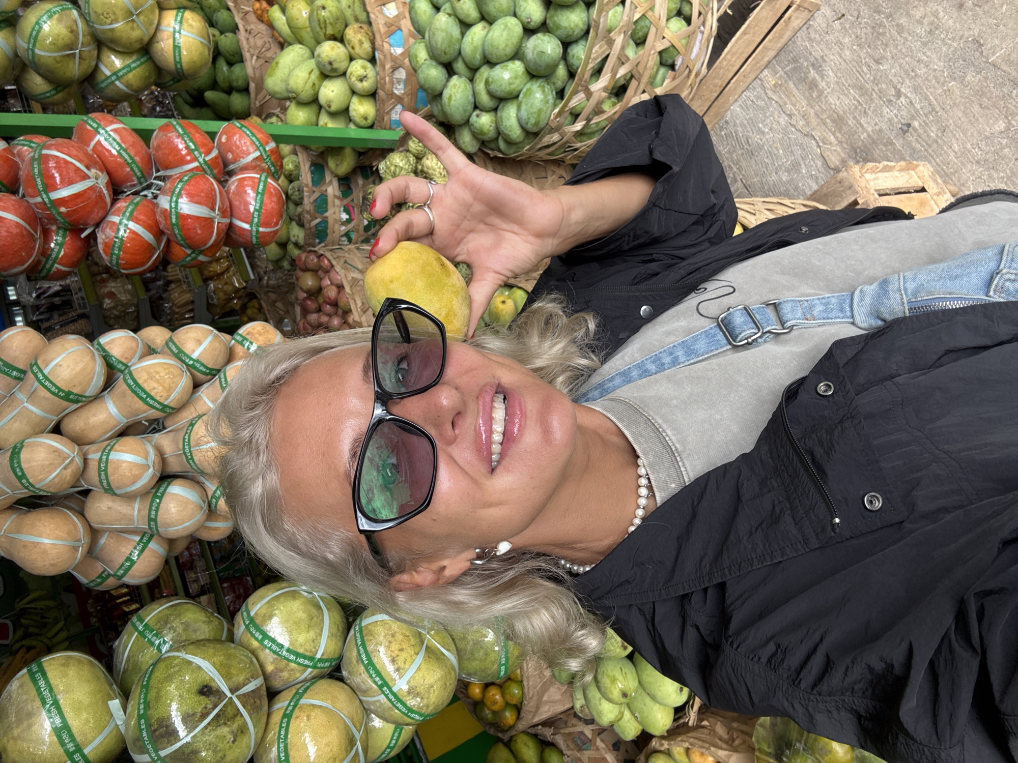 The image shows a woman holding a yellow fruit, likely a mango, in front of a display of produce at a market. She has silver hair, is wearing sunglasses and a black jacket over a gray shirt, and is smiling. A pearl necklace is visible around her neck, and she has several rings on her fingers. The produce in the background includes items wrapped in clear plastic with labels in an Asian language, such as mandarin oranges and other fruits or vegetables. There are also unwrapped fruits such as green mangoes. The woman is either shopping or taking a picture of herself with the market display in the background. The location is Unknown, Unknown.