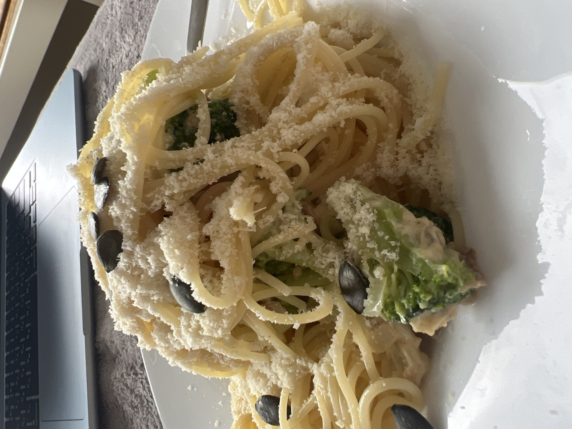 A plate of spaghetti with broccoli, shredded cheese, and black olives sits on a white plate next to a laptop. The scene appears to be indoors, possibly in an apartment or hotel room. The location is unknown, so the city of Budva cannot be confirmed.