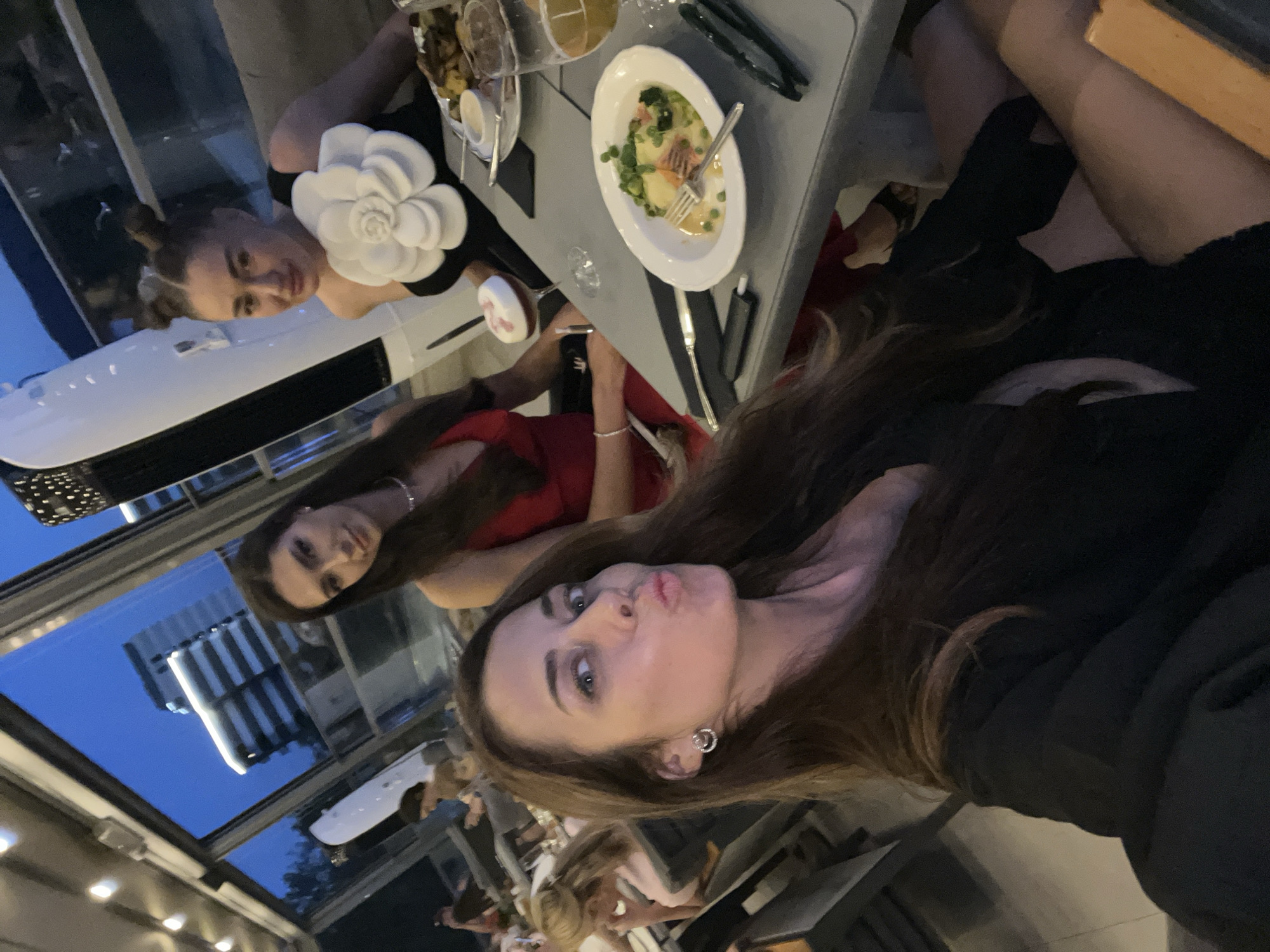 A selfie taken at an outdoor dining establishment in Boreti, Montenegro, shows three women seated at a table. The woman in the foreground is looking directly at the camera, puckering her lips. She has long, dark brown hair and is wearing a black top and earrings. To her left, a woman in a red dress is visible, looking forward. To the right of the foreground woman, another woman with her hair styled in two buns is smiling. She is wearing a black top adorned with a large white flower. On the table in front of them are plates of food, drinks, and cutlery. The background shows a view of buildings and the sky as dusk settles.