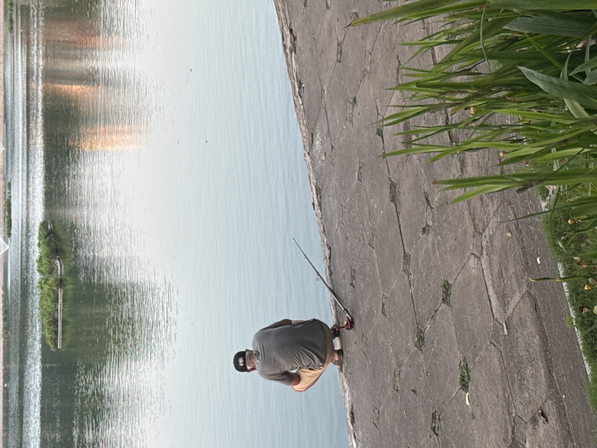 The image depicts a man fishing in Hoan Kiem Lake in Hanoi, Vietnam. The man is kneeling on a concrete walkway, facing the lake. He is wearing a grey shirt, khaki shorts, sandals, and a black baseball cap. A fishing rod rests on the walkway next to him. The lake is visible in the background, with reflections of the sky and buildings visible on the water's surface. The edge of the walkway is lined with green plants.