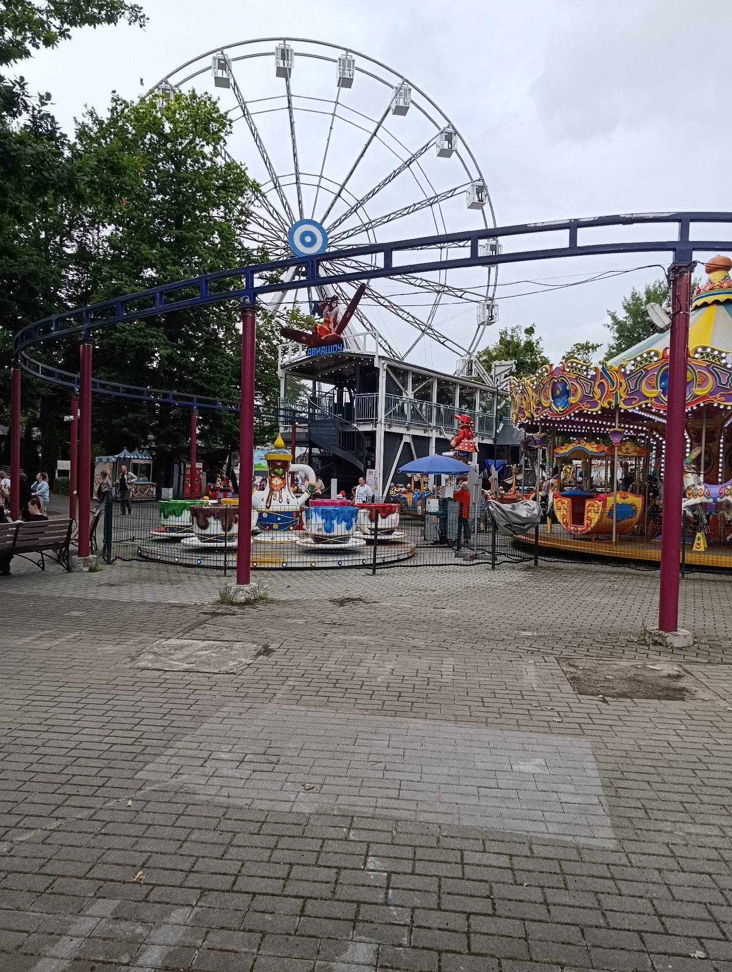The image captures an amusement park scene, likely within Kaliningrad, Russia, given the visual cues. Dominating the background is a large Ferris wheel with white cabins against a cloudy sky. In the middle ground, a colorful carousel with ornate decorations is visible, alongside a children's tea-cup ride with cups painted in blue, white, and brown. A roller coaster track snakes through the scene, supported by blue and red poles. Various people are scattered throughout the park, some seated on a wooden bench in the foreground, others standing near the rides. The ground is paved with gray bricks. The overall atmosphere suggests a typical day at a local amusement park.