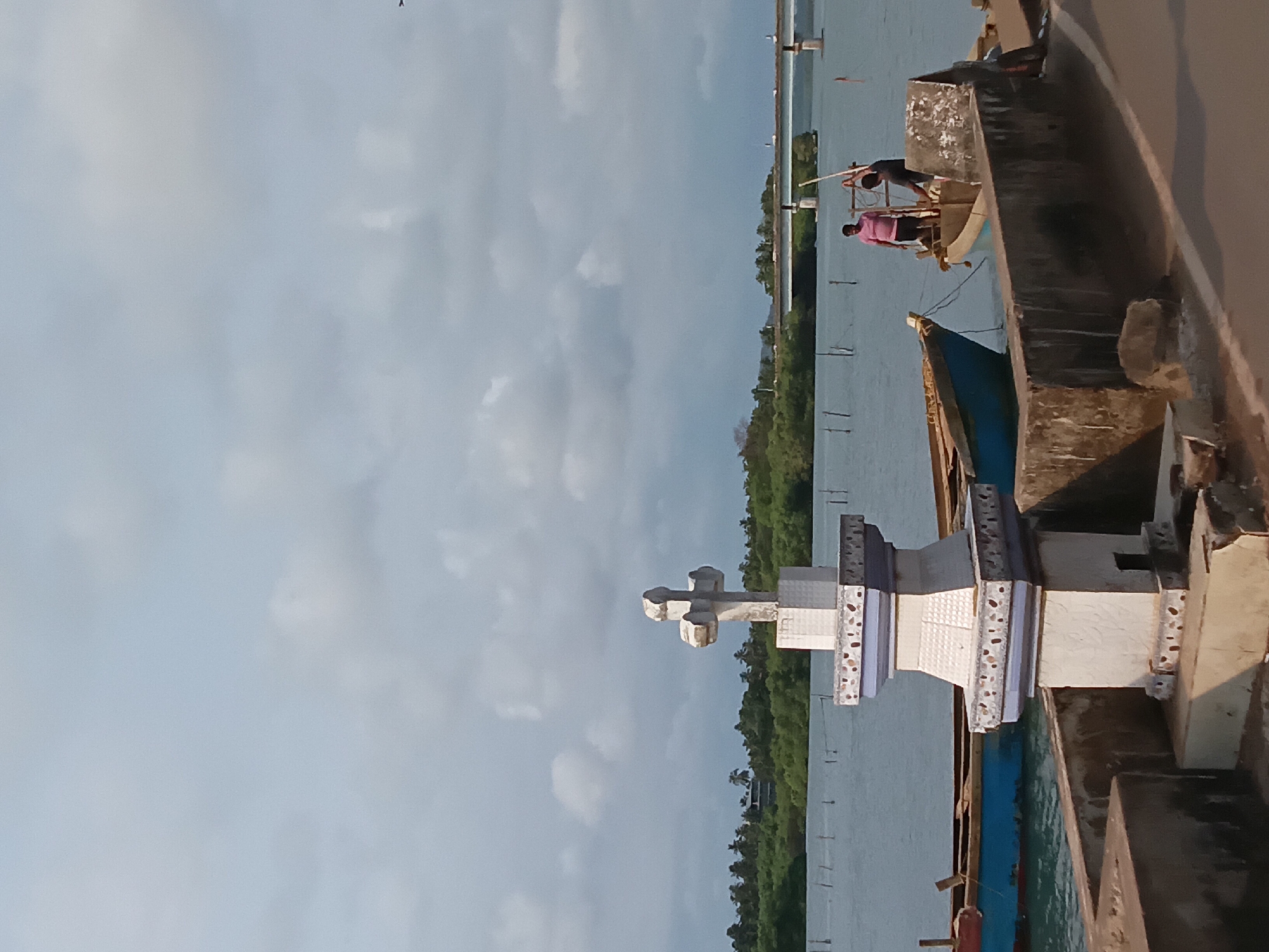 Here's a description of the image:
The image shows a waterfront scene in Oxel, India. A concrete cross monument stands prominently in the foreground. Beside the monument, two men are visible in a boat; one is wearing a pink shirt and the other is wearing a dark shirt. Both men are using long sticks to propel the boat. The water extends behind the boat, with a dense line of green trees visible on the opposite bank. A bridge is faintly visible further in the background. The sky above is light blue with scattered clouds. The scene is viewed from a slightly elevated position, likely from a bridge or dock.