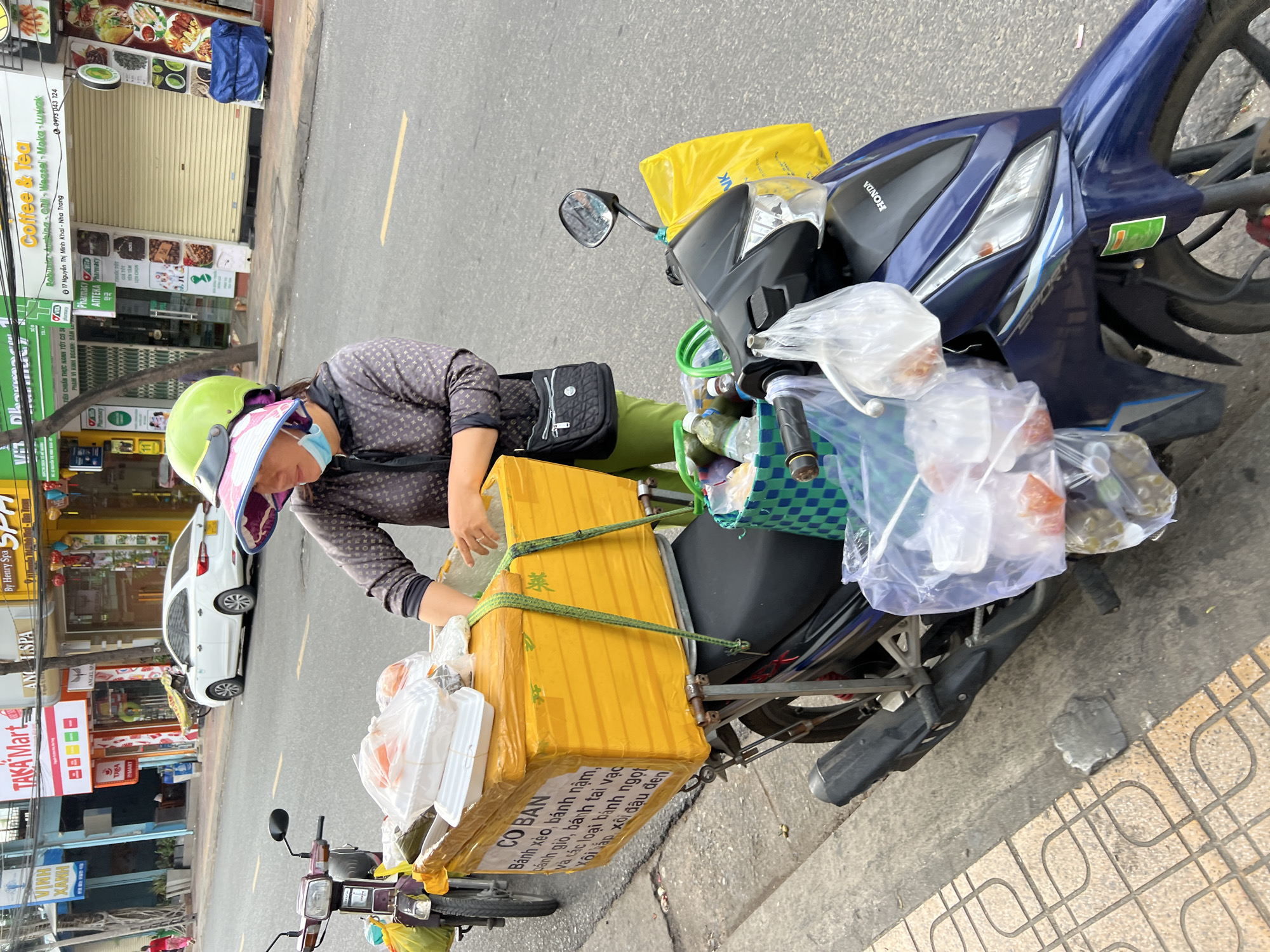 A woman wearing a green helmet, a face mask, and a patterned shirt stands beside a blue motorcycle parked on the side of a street in Nha Trang, Vietnam. She is reaching into a large, yellow cooler box attached to the back of the motorcycle. The cooler box is strapped on and contains several white food containers and plastic bags filled with food items. A green and blue checkered basket is also attached to the motorcycle, holding bottles of drinks. The street is paved with asphalt, and there are buildings with shops on the right side of the image. The shops appear to be a Taka Mart, a pharmacy, and a coffee and tea shop. A white car is parked further down the street. The overall scene depicts a street vendor preparing to sell food from her motorcycle.