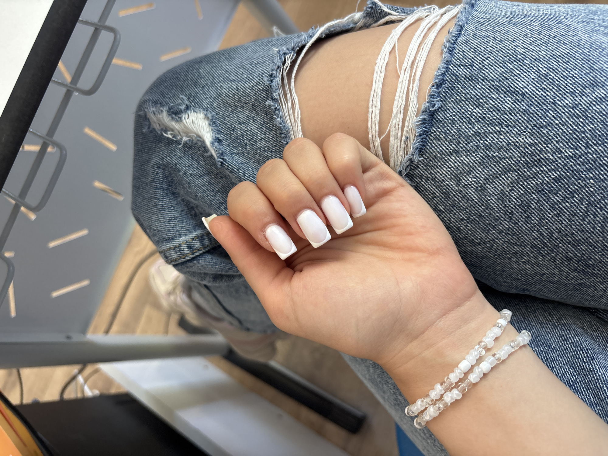 A woman's hand, adorned with white French tip acrylic nails and a delicate beaded bracelet, rests on her lap, which is covered by ripped denim jeans. The background shows a partial view of a desk and chair, suggesting an indoor setting such as a home or office. The scene is well-lit, highlighting the details of the woman's hand and attire. There is no identifiable information about the location in Shumen, Bulgaria.