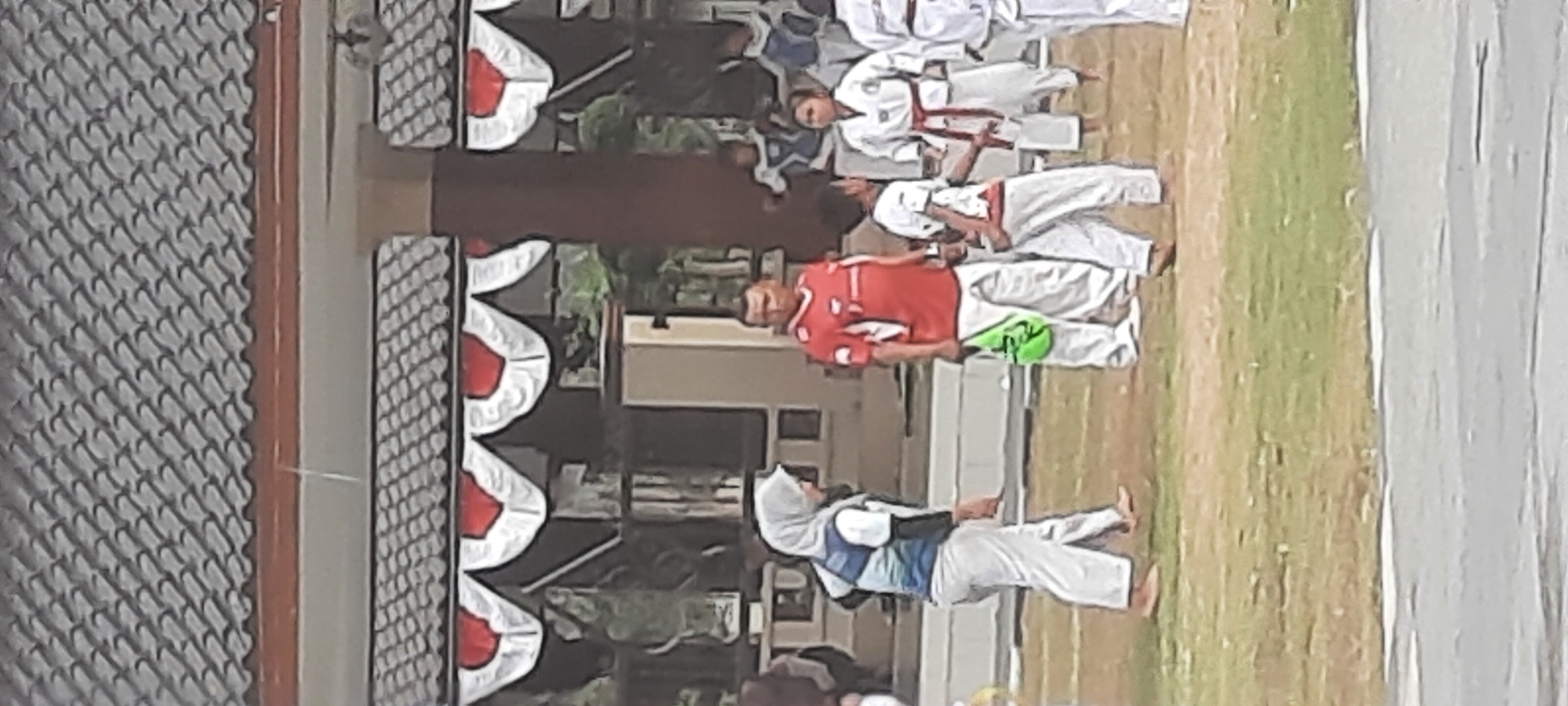 The image shows an outdoor scene likely in Kanigoro, Indonesia, with several people gathered on a grassy area in front of a building with a distinctive tiled roof and red and white decorative banners. In the foreground, a woman wearing a hijab and a martial arts uniform is walking. To her right, a man in a red shirt and white pants stands holding a green bag. Behind them, several children in white martial arts uniforms are visible, some with belts. The environment appears to be a public space, possibly a schoolyard or community center, given the presence of the building and the organized gathering. The lighting suggests it is daytime.