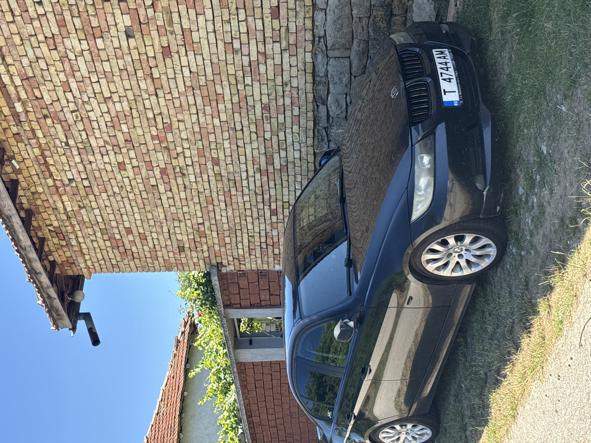 A black BMW sedan is parked on grass in front of a brick building in Konop, Bulgaria. The building has a red-tiled roof and a rough stone foundation. The car's license plate is T 4744 AM. The sky is clear and blue.