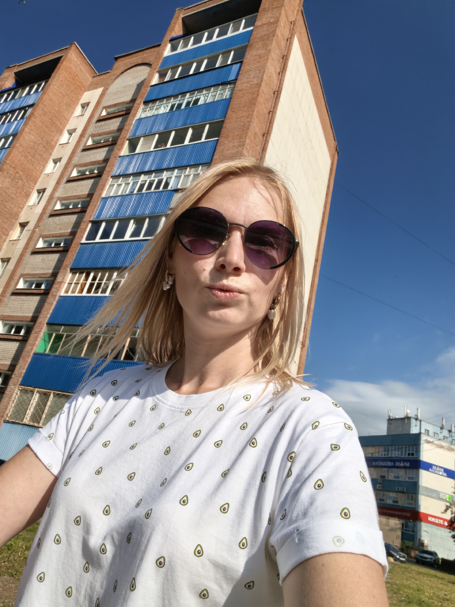 A young Caucasian woman with blonde hair and sunglasses takes a selfie outdoors in front of a multi-story brick building with blue balconies. She is wearing a white t-shirt with a pattern of small avocados. The building appears to be in an urban environment. The sky is clear and blue. The image is likely taken in the city of Sterlitamak, Russia.