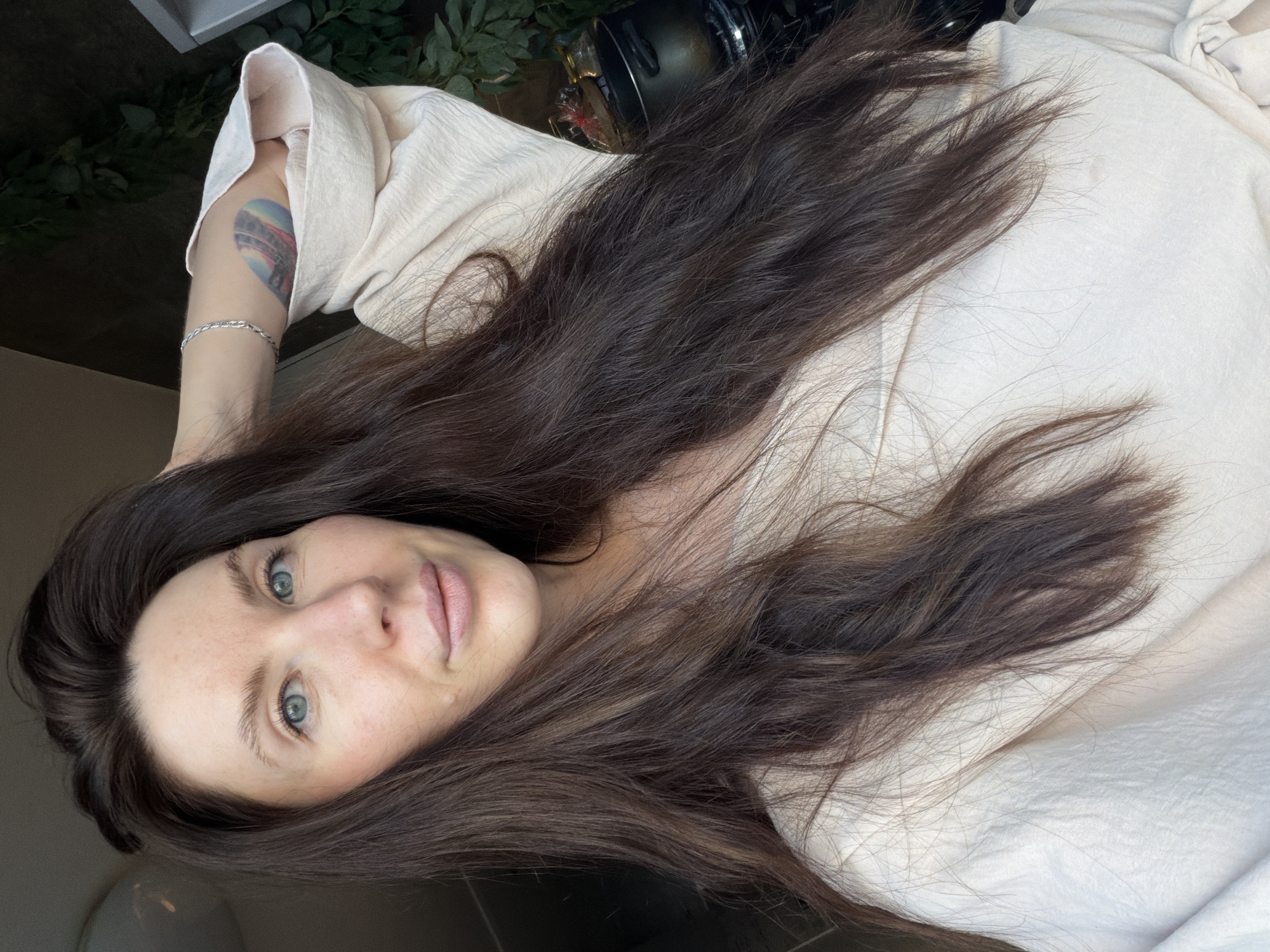 A medium-shot, portrait photograph taken indoors, depicts a woman with long, dark brown hair lying on what appears to be a white fabric or bedsheet. Her head is positioned in the lower-left corner of the frame, angled towards the camera, revealing her face with fair skin, light blue eyes, and light pink lips. The woman is wearing a light-colored, possibly off-white or beige, garment with a visible sleeve draped over her arm, which also displays a tattoo on the upper arm and a silver bracelet on the wrist. The background shows blurred elements, including what appears to be greenery and a dark cylindrical object, possibly a camera lens. The photograph is taken from a slightly elevated perspective, with the focus primarily on the woman's face and hair. There is no visible indication of the location being in Perm, Russia.