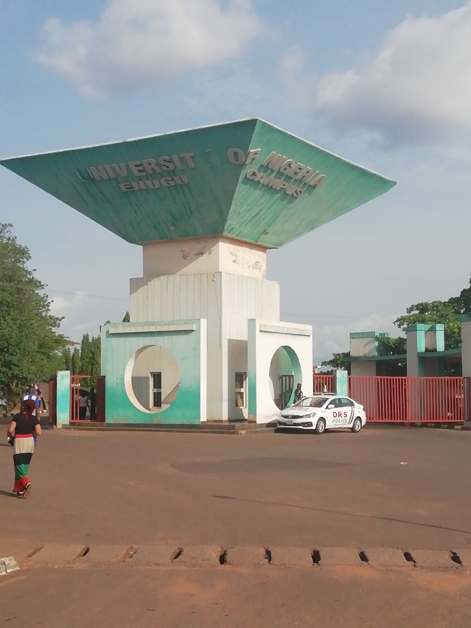 The image depicts the entrance gate to the University of Nigeria, Enugu Campus, located in Enugu, Nigeria. A large, square-shaped structure painted green and white sits atop the gate, bearing the inscription "UNIVERSITY OF NIGERIA ENUGU CAMPUS". Two circular openings are present on either side of the central structure. A white police car with "DRS POLICE" marked on its side is parked in front of the gate. Several people are visible in the background entering and exiting the gate. A person is walking in the foreground, away from the camera. The road in front of the gate is reddish-brown. The sky is partly cloudy. Trees and greenery are present in the background.