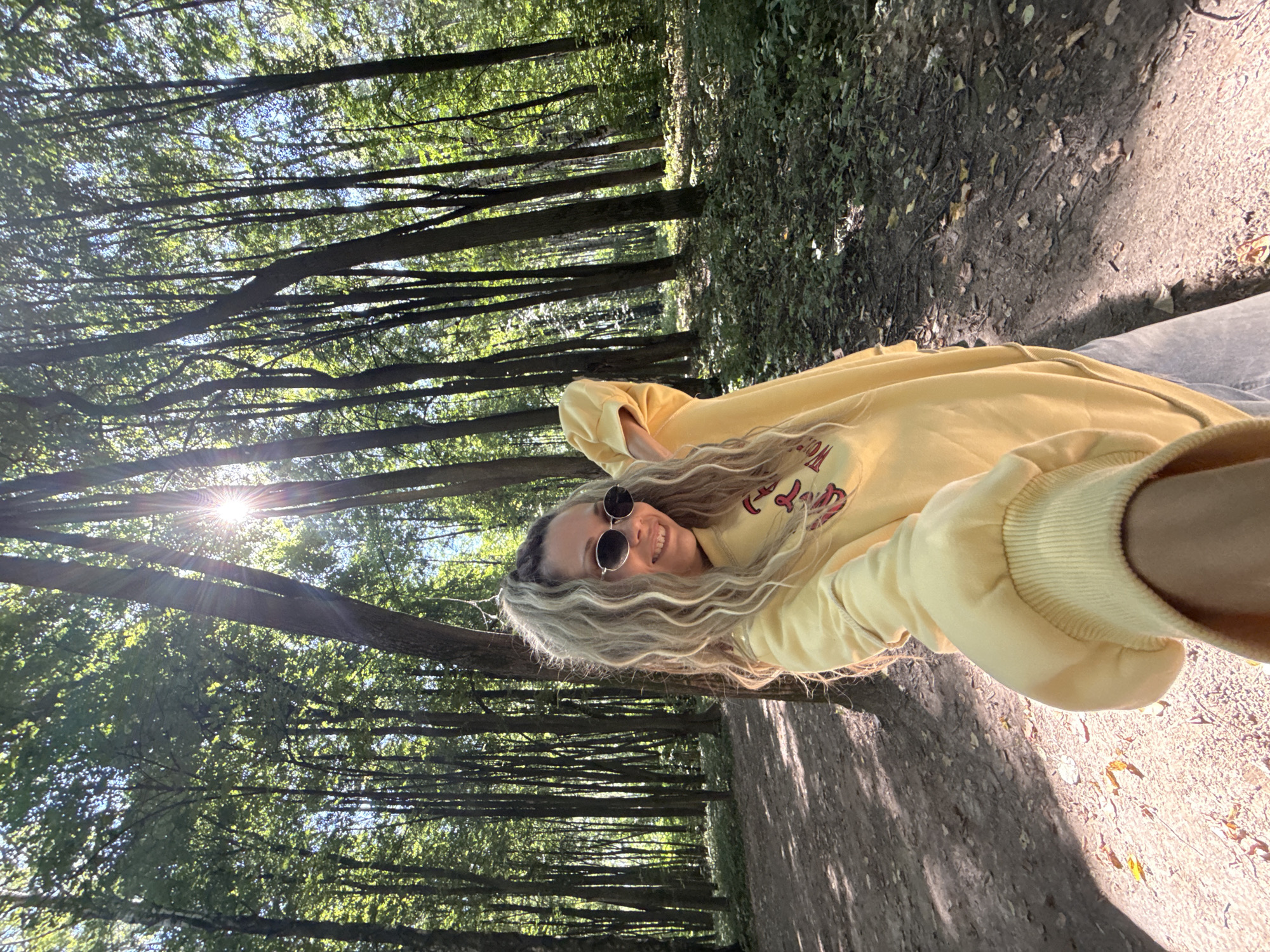 A young woman with long, wavy blonde hair and sunglasses smiles at the camera, holding out her arm and the camera. She is wearing a yellow sweatshirt and light-colored pants. She stands in a forest with tall trees and dappled sunlight. The ground is covered with dirt, leaves, and some patches of grass. The scene is likely in the city of Rusanovo, Russia.
