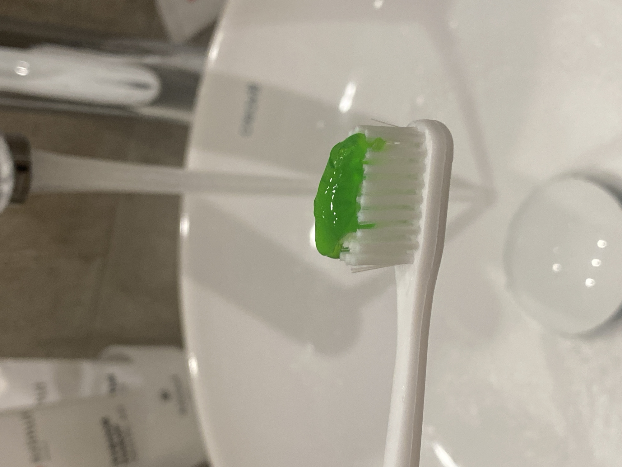 A white toothbrush with green toothpaste sits atop a white bathroom sink. The sink has a silver drain cover and a faucet dispensing water. In the background, partially visible, are toiletries and a section of a tiled wall. The location in the city of Sochi, Russia is not determinable from the image.